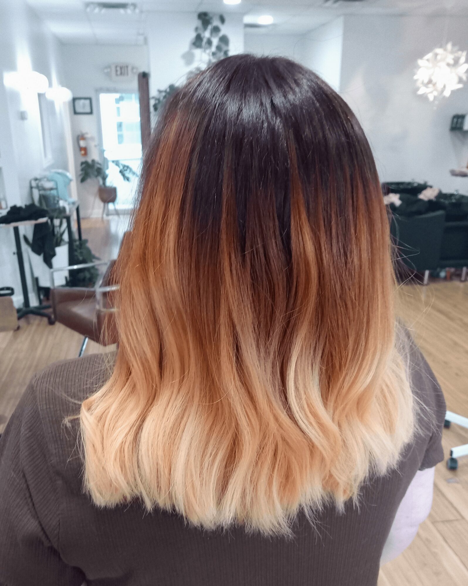 Back of a person's head with long, wavy hair transitioning from dark brown to blonde, in a salon setting.