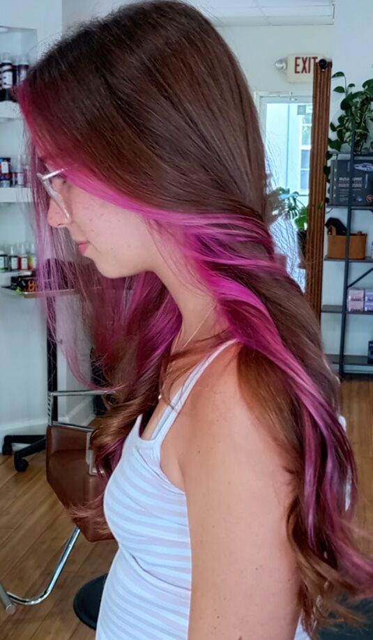 Side view of a woman with long, wavy hair dyed pink and purple, wearing glasses and a striped tank top.