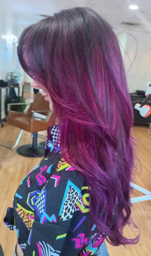 Side view of a person with long, wavy hair dyed in shades of purple and pink, in a salon setting.