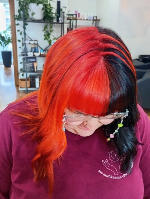 Person with half red and black hair, wearing glasses, looking downward in an indoor setting.