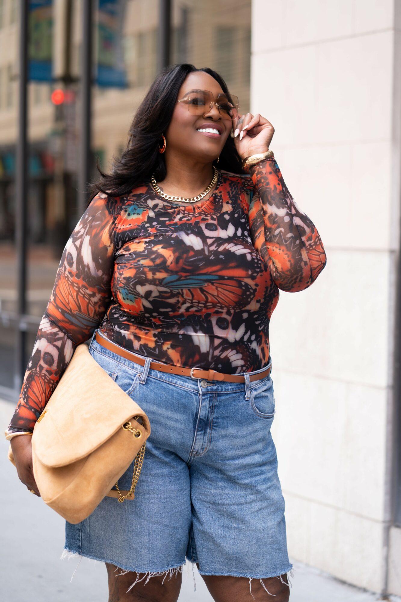 Woman smiling and talking on phone, wearing patterned top and denim shorts, carrying a tan bag, standing outdoors.