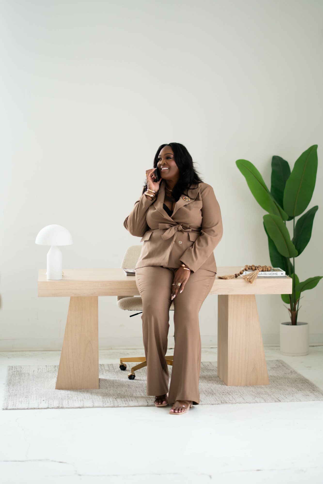Woman in beige outfit sitting on a chair, talking on phone, in a minimalist room with a large plant and desk.