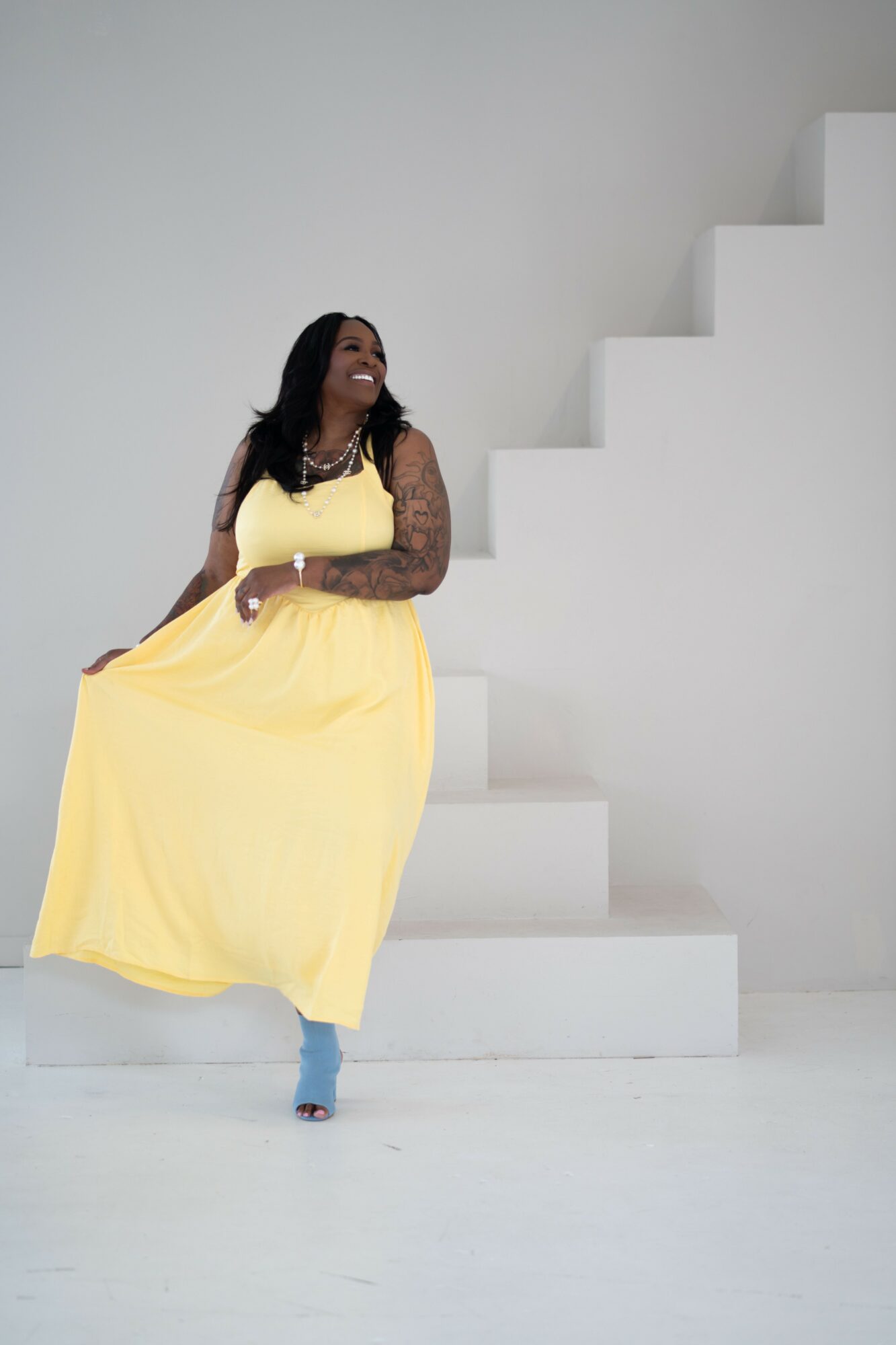 Woman in yellow dress sitting on stairs, smiling, against a plain white background.