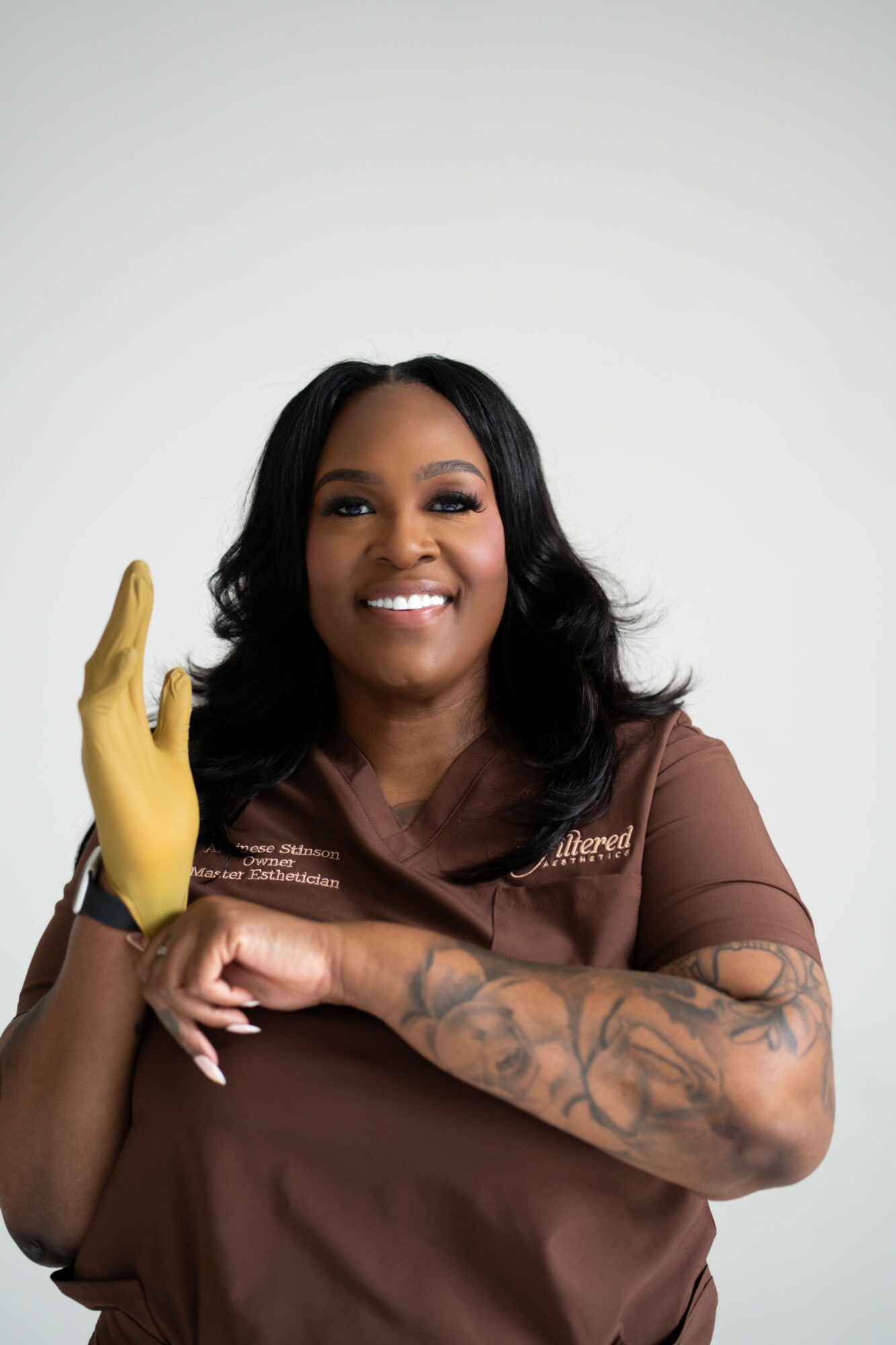 Smiling woman with black hair and tattoos on her arm, wearing a brown shirt and yellow glove, against a plain background.