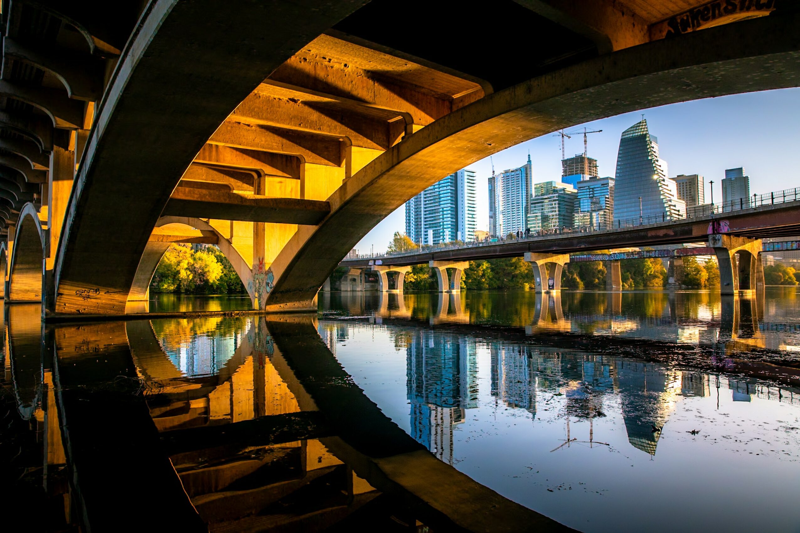 Under a bridge with city skyline and water reflection, sunlight illuminates the scene, showing modern buildings and a clear sky.