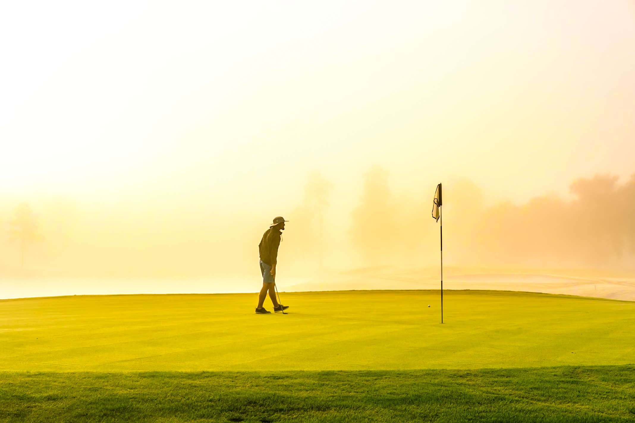 Person standing on a golf course near a flagstick, with a bright sky in the background.