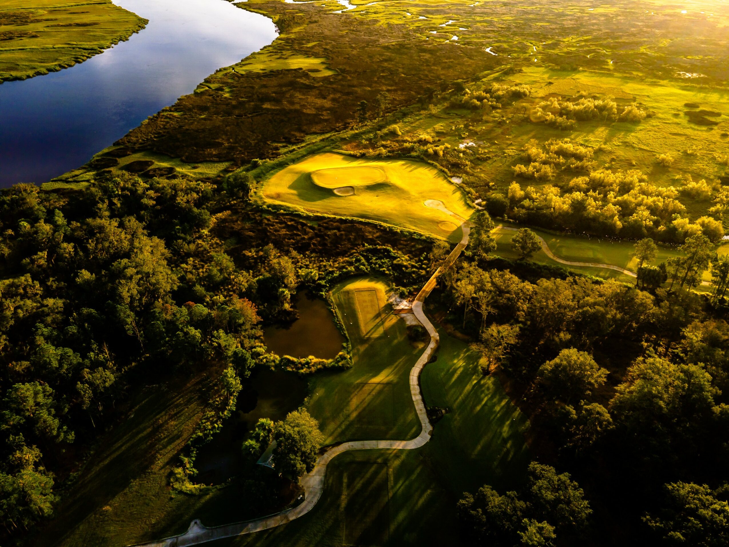 Aerial view of a river, sand dunes, and green landscape with a winding road, trees, and open fields.