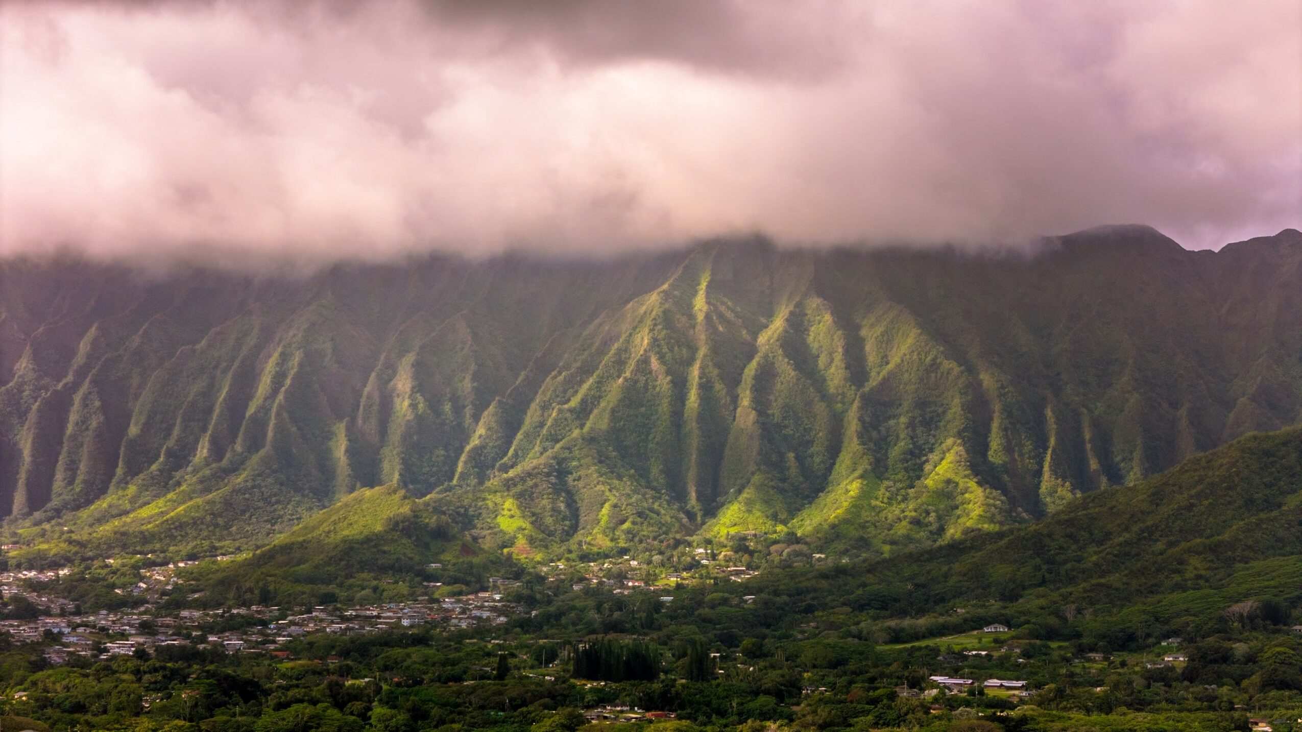 Mountain range with green slopes and a cloudy sky, overlooking a valley with buildings.