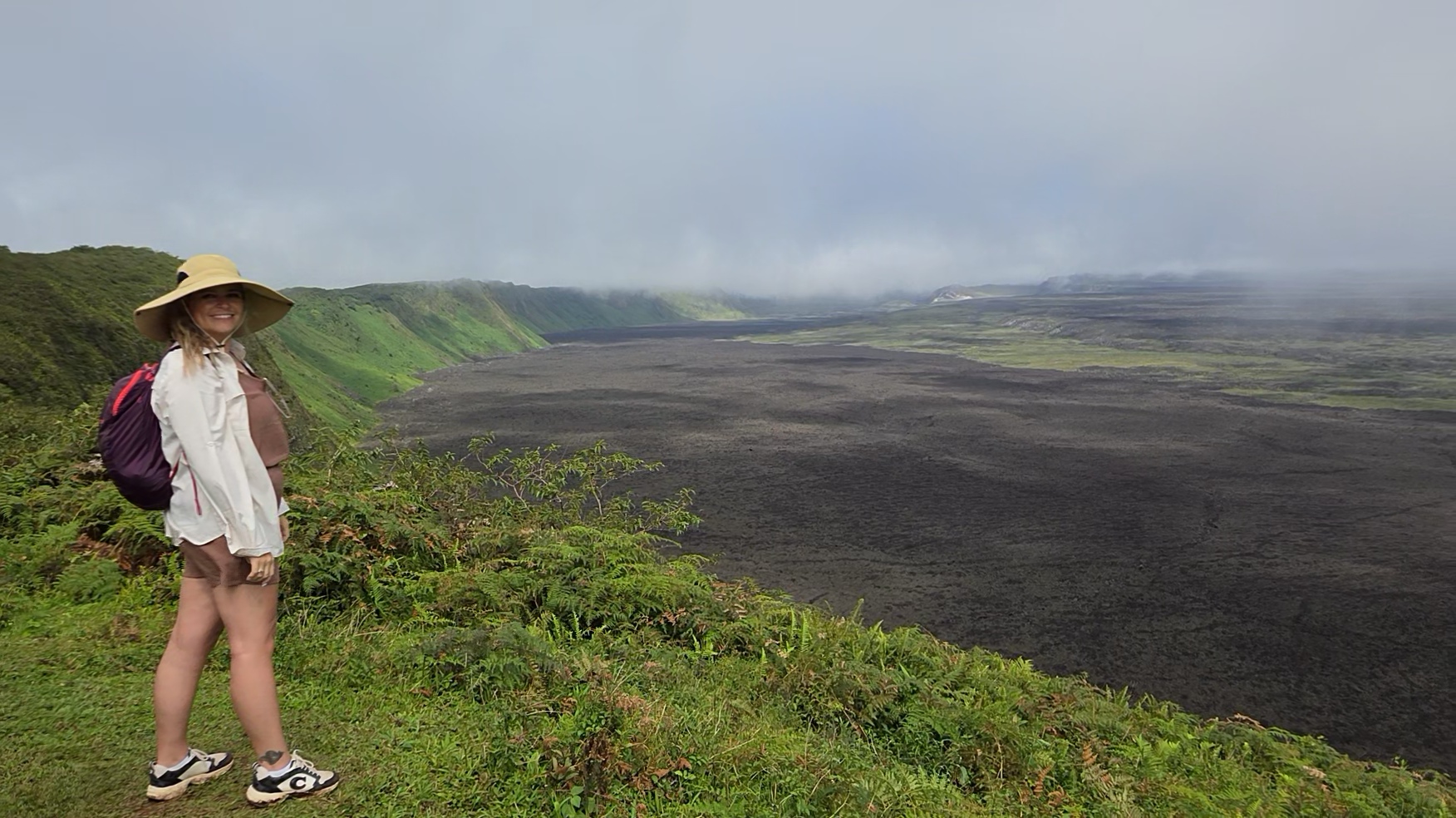 Girl wearing a hat and backpack standing on grassy hill overlooking volcanic landscape with clouds above.