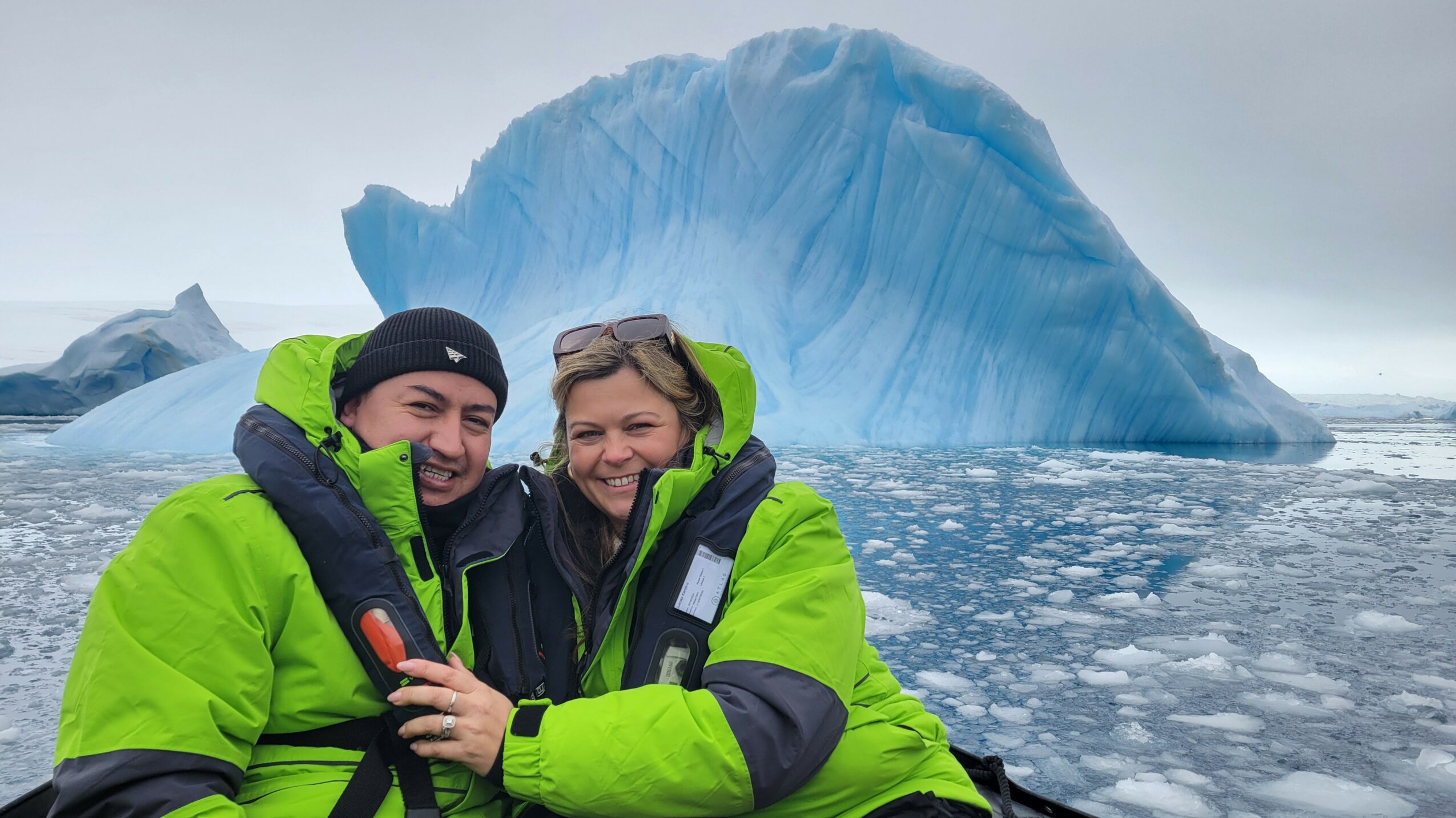 Two people in bright green jackets smiling in front of a large iceberg in icy water.