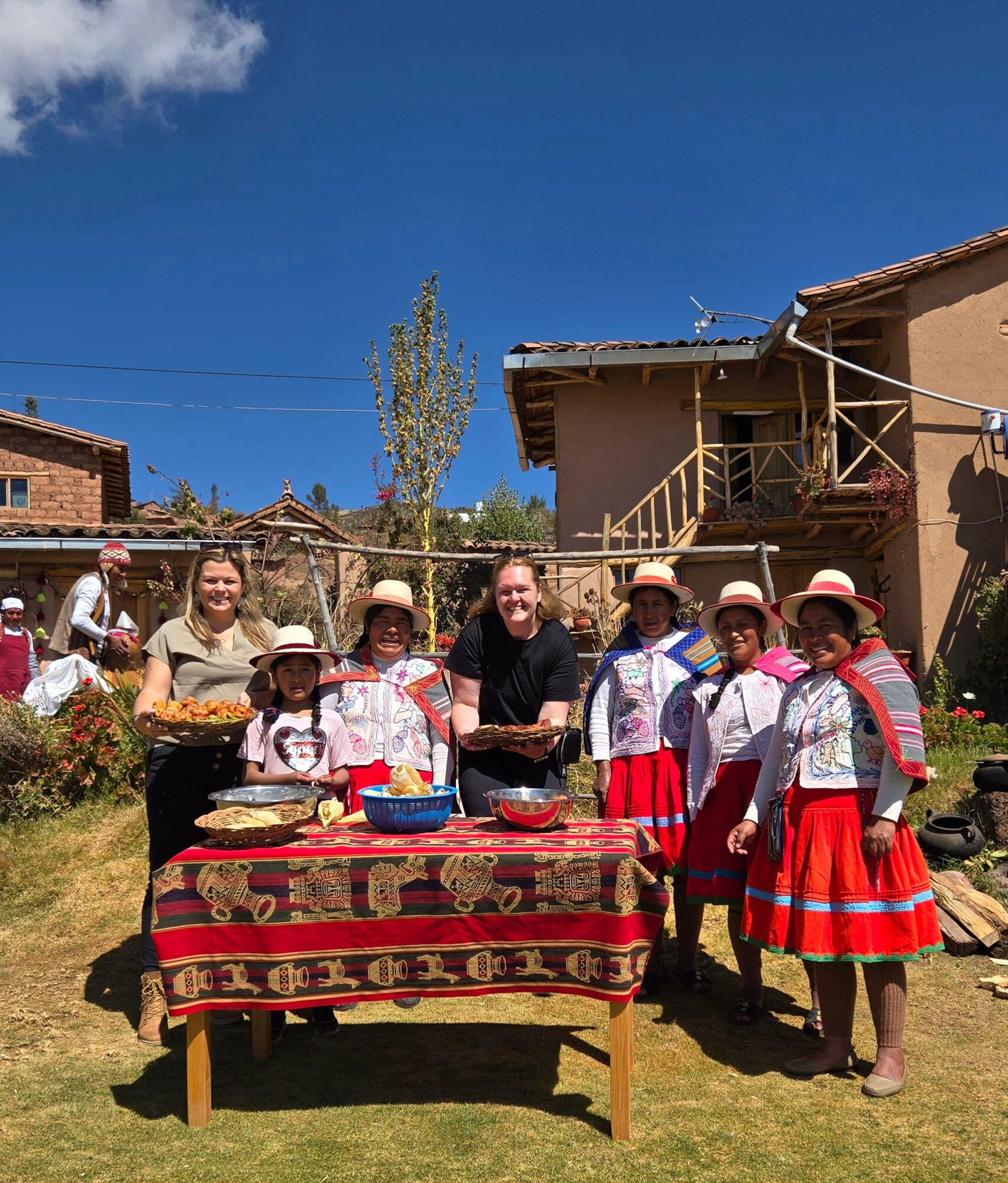 Group of people in traditional clothing around a table outdoors, with a house and blue sky in background.