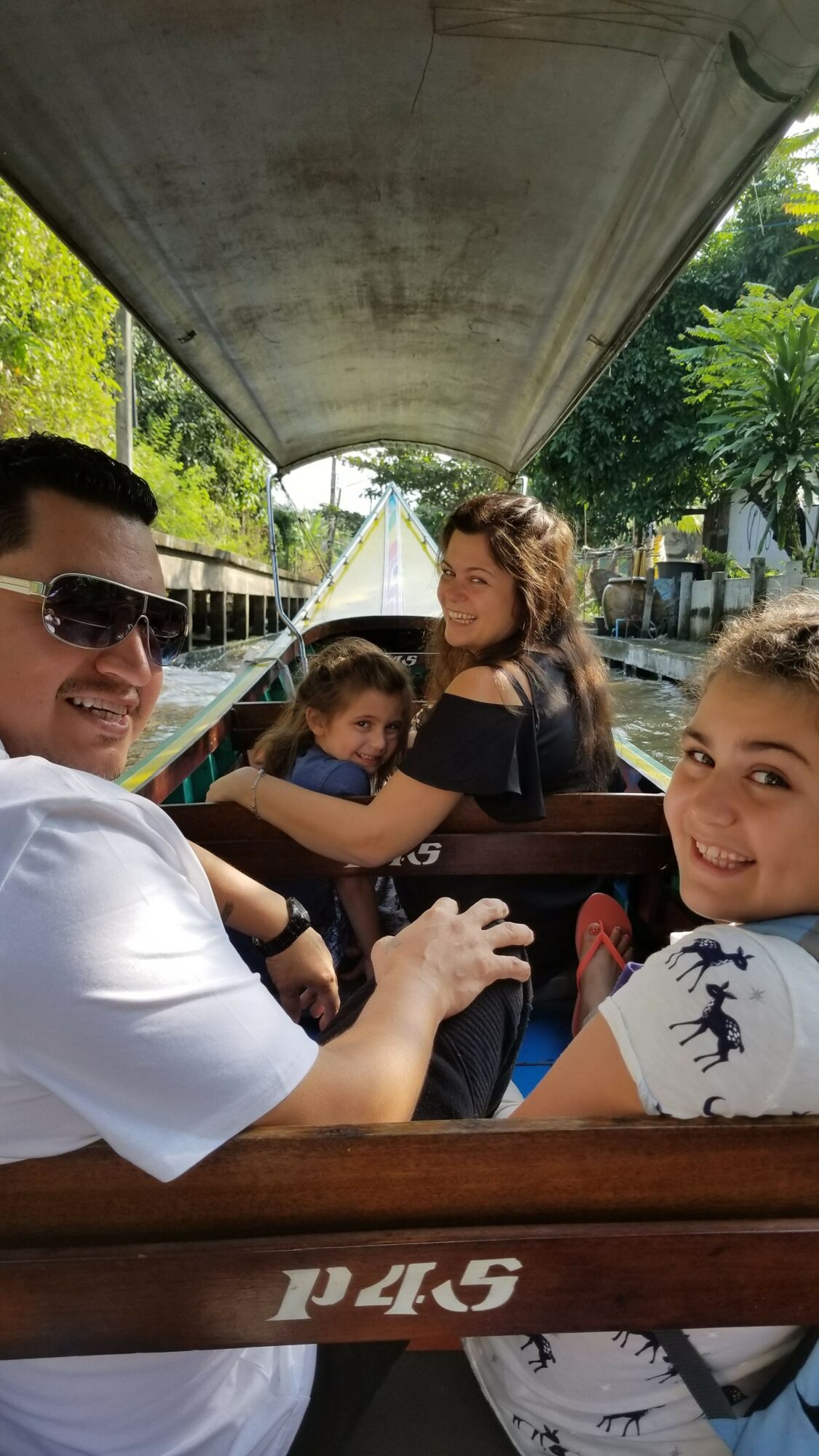 Group of children and an adult sitting at a wooden table outdoors, with a large concrete structure overhead and trees in the background.