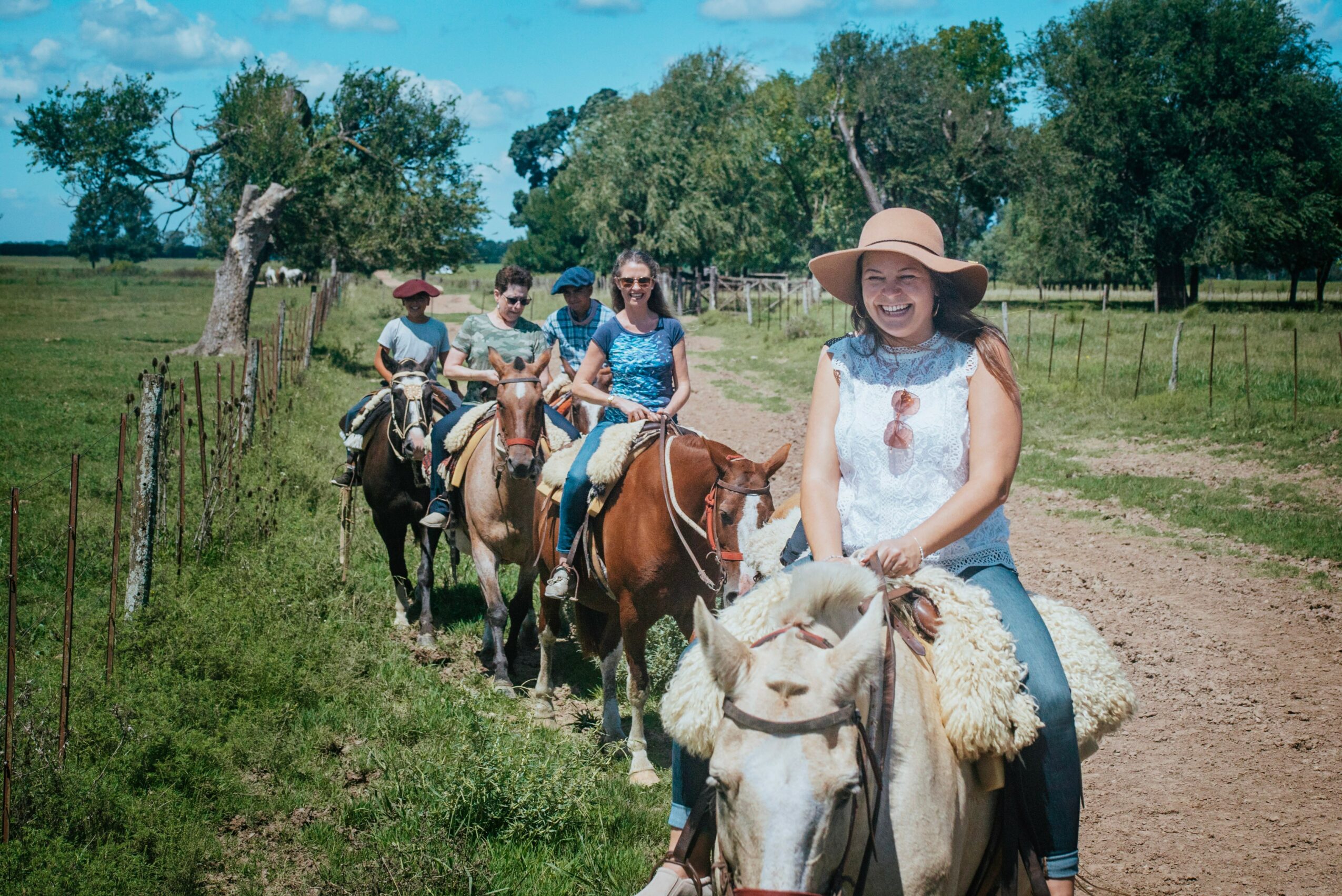Group of four people riding horses on a dirt path in a grassy field with trees, sunny weather, and blue sky.
