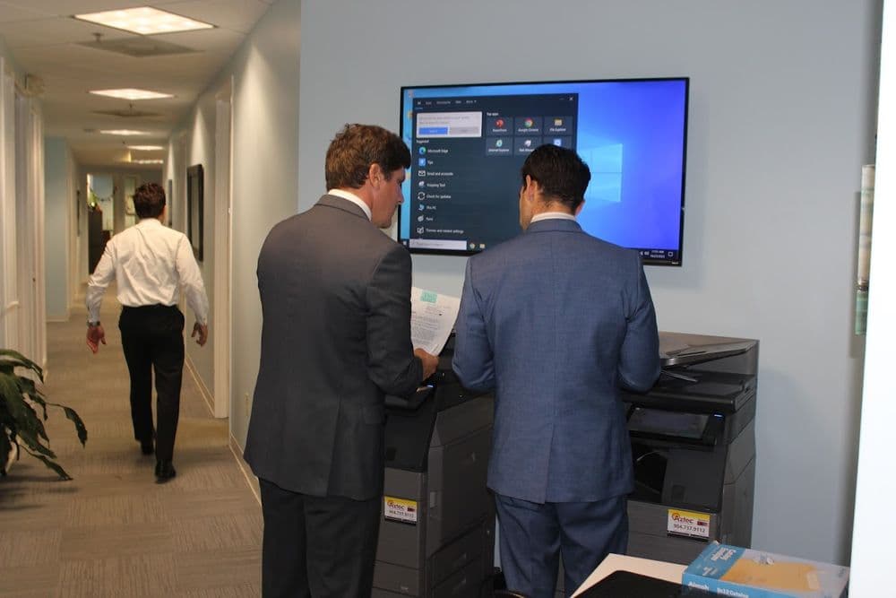 Two men in suits using a touchscreen kiosk in an office hallway, with a person walking in the background.