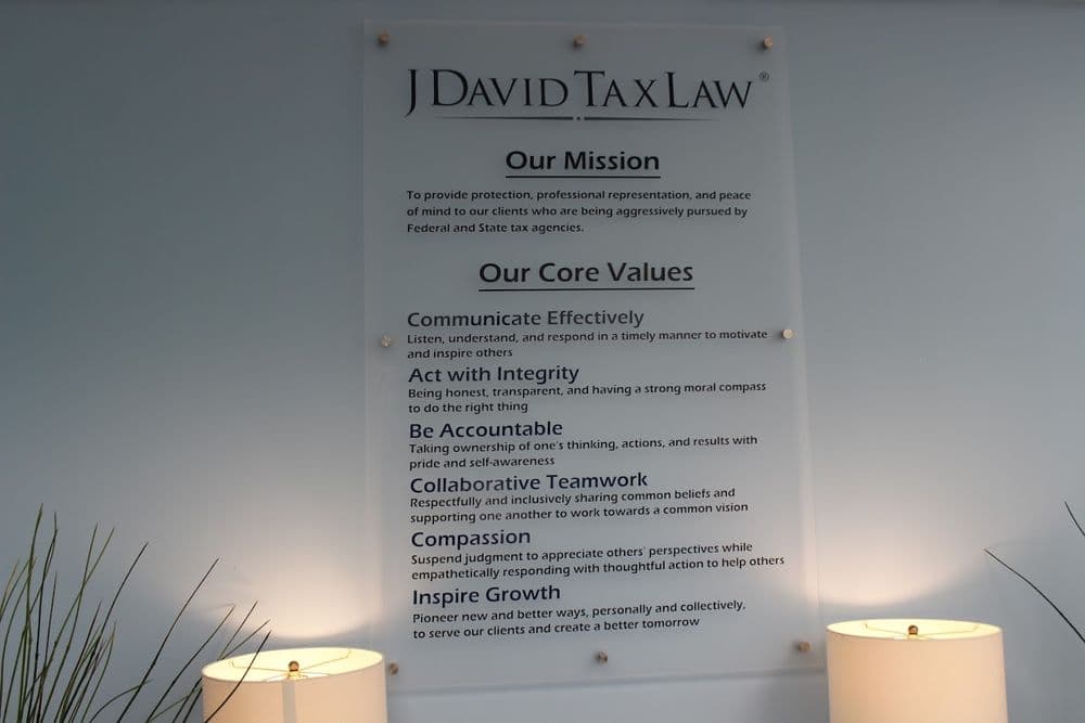 A poster titled 'J David Tax Law' lists mission and values, with two lamps at the bottom, against a light-colored wall.