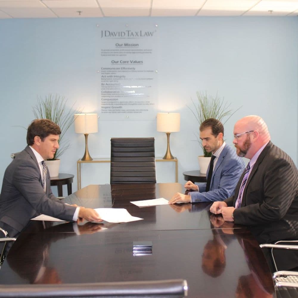 Three men in suits sit at a conference table in a room with a blue wall, two lamps, and plants, discussing documents.