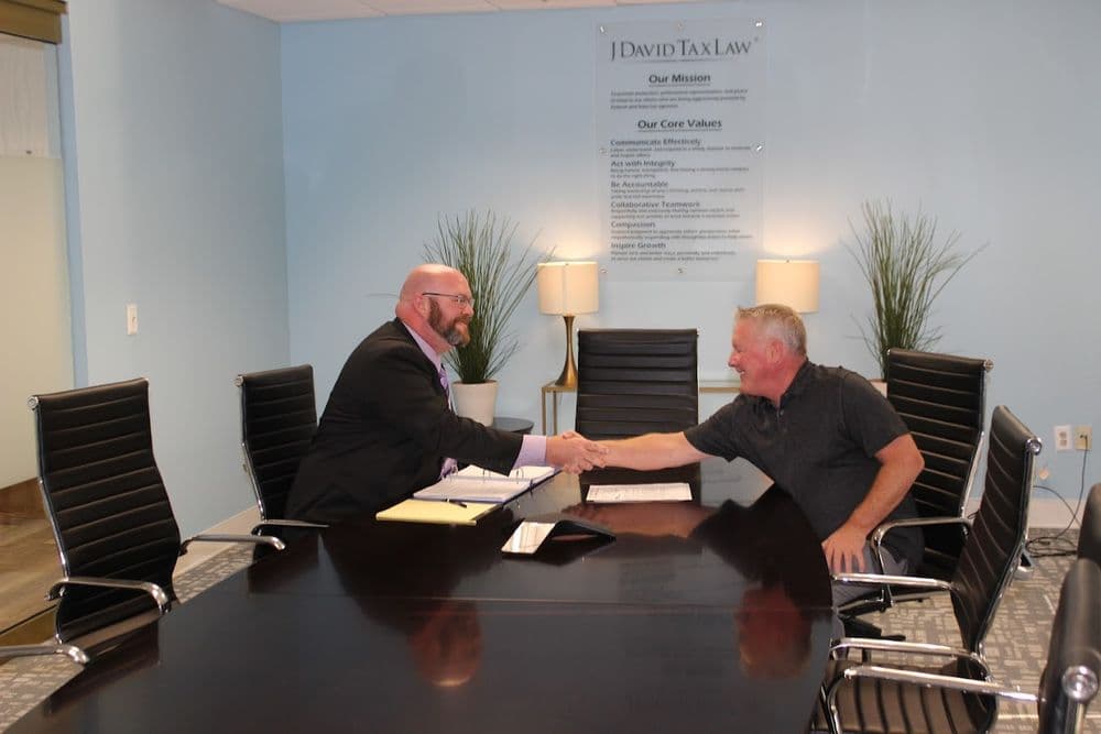 Two men shake hands across a conference table in a room with blue walls, plants, and a poster on the wall.