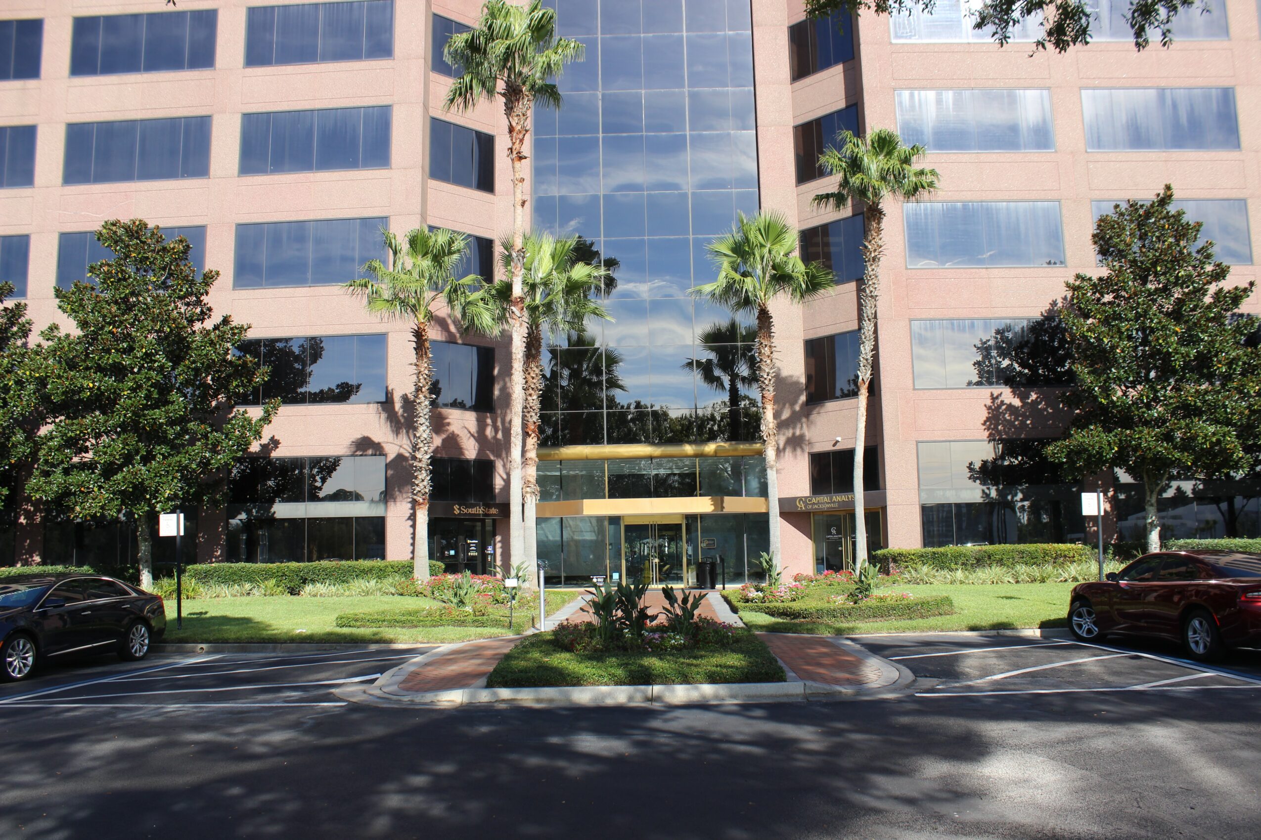 Modern office building with glass windows, surrounded by palm trees and greenery, with parked cars in front.