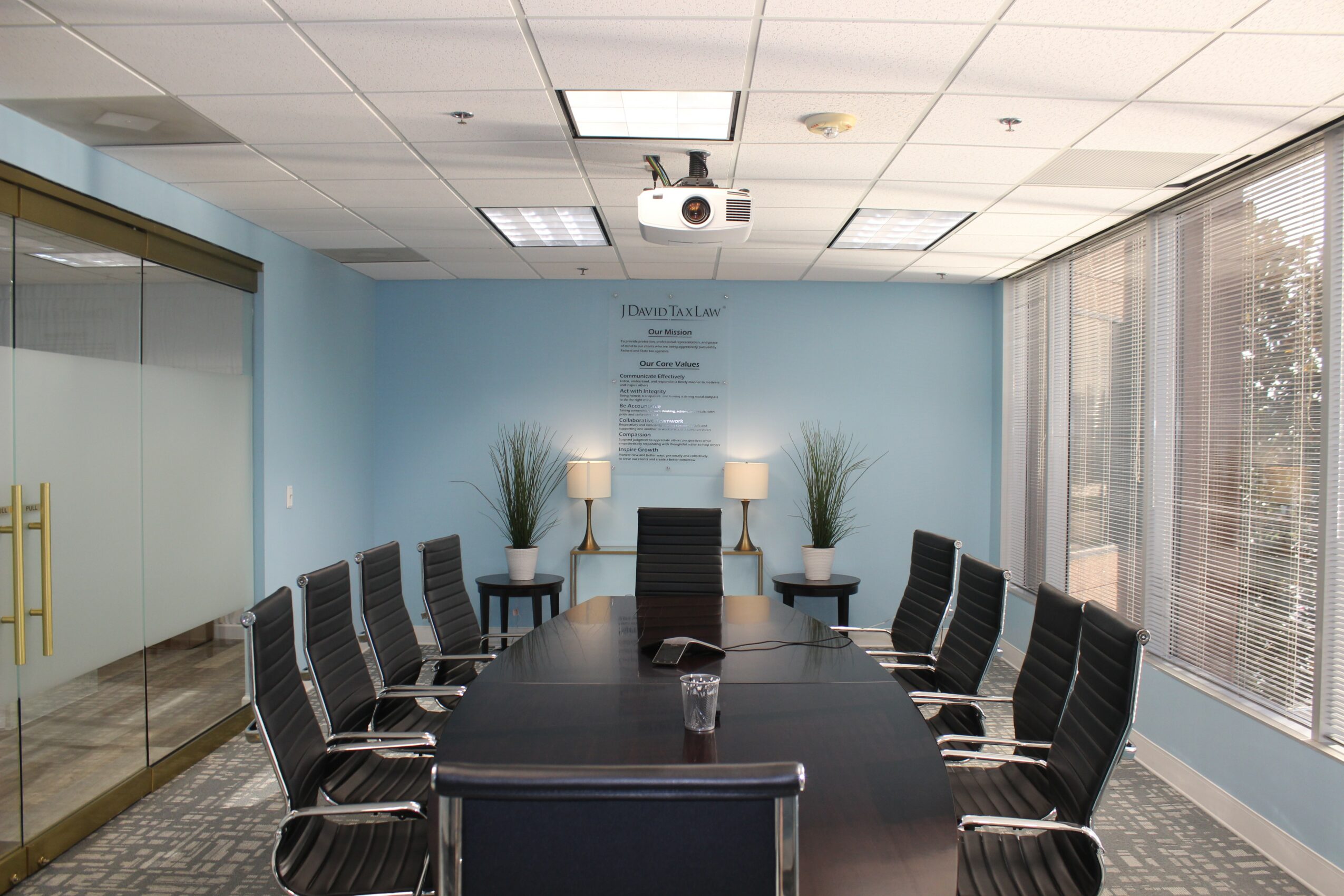 Conference room with a long table, ten chairs, two potted plants, and large windows on the right side.