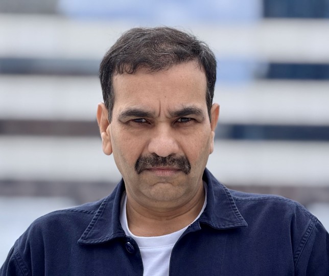 Man with dark hair and mustache wearing a dark jacket and white shirt, outdoors with blurred background.