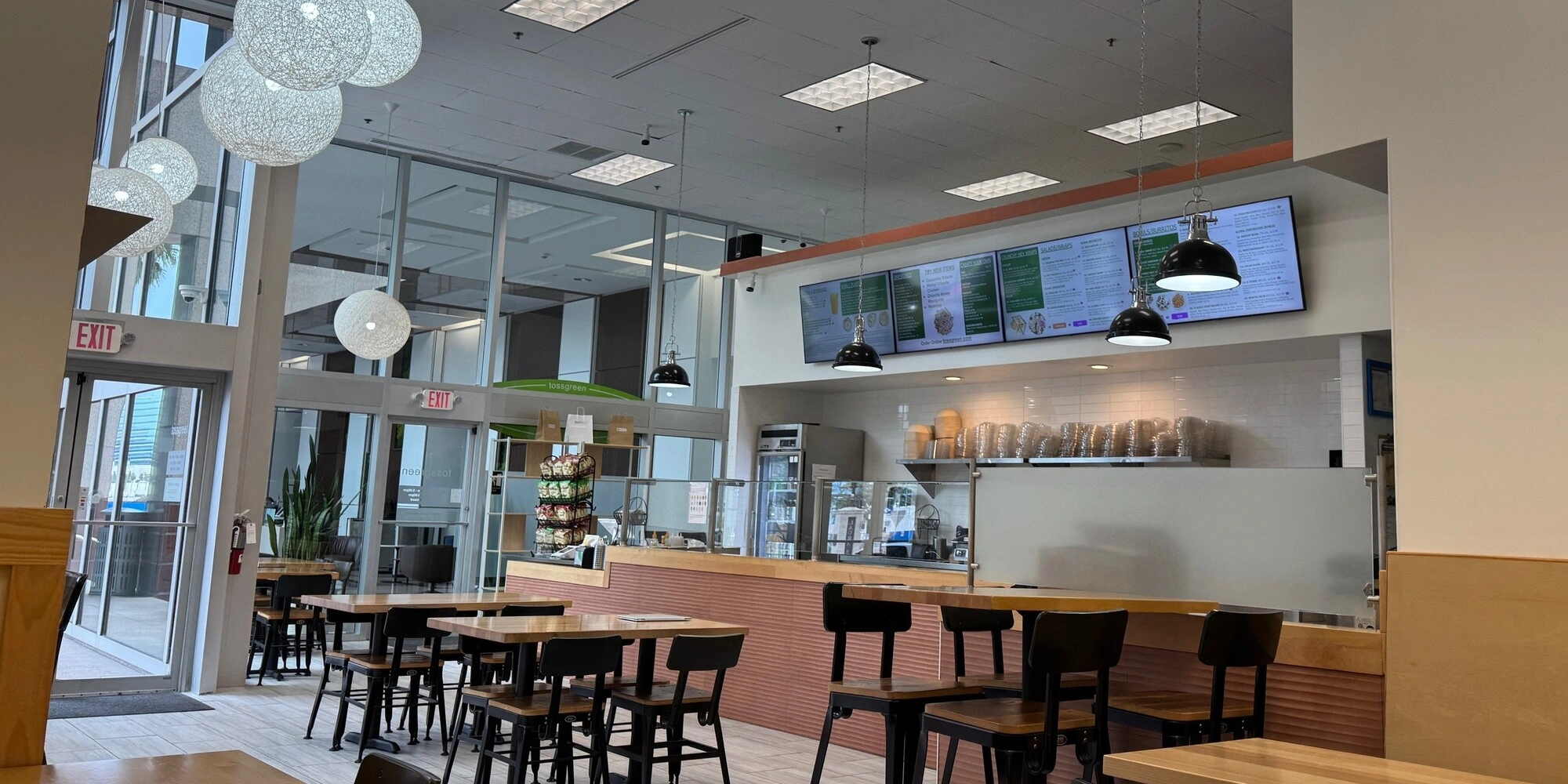 Interior of a modern cafe with tables, chairs, large windows, and digital screens on the wall.
