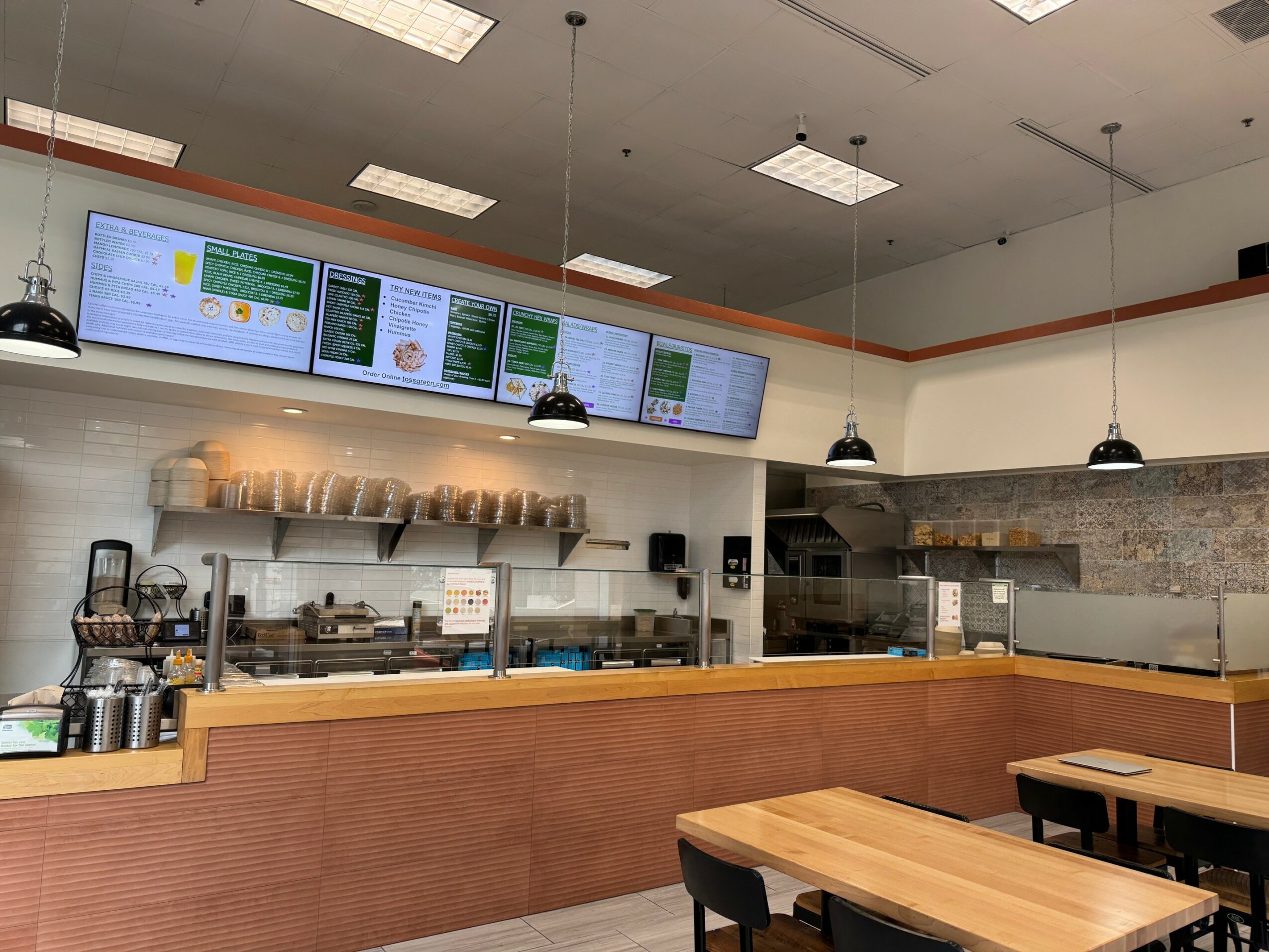 Interior of a cafe with digital menu screens, wooden tables, and a counter area with equipment and supplies.