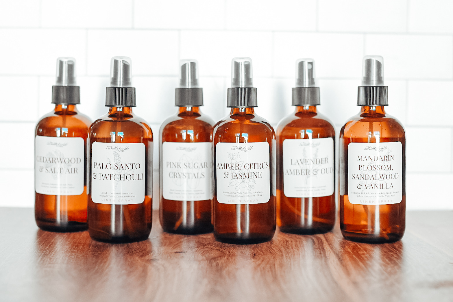 Six amber spray bottles with white labels and gray caps on a wooden surface, against a white tiled background.