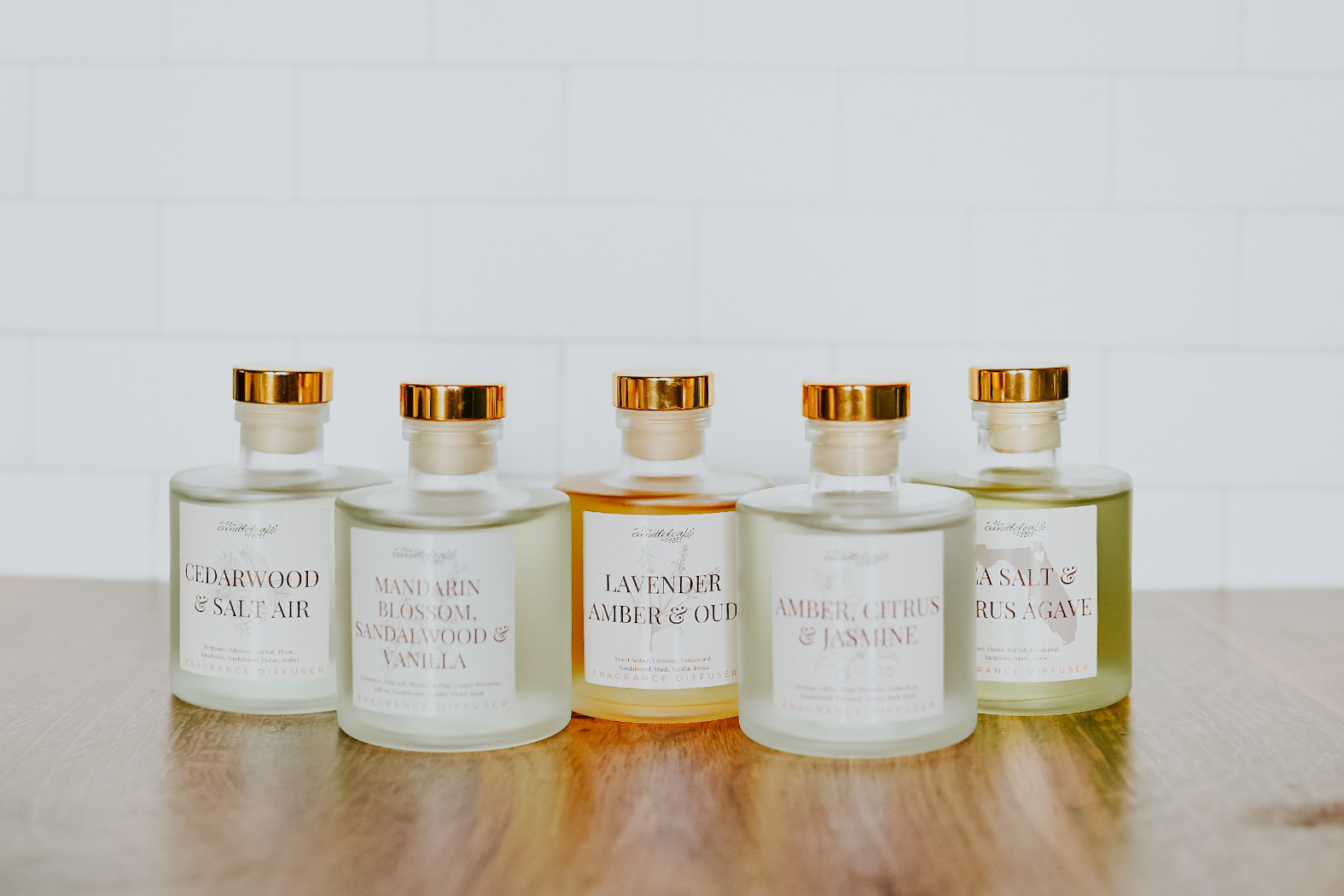 Five glass bottles with gold caps containing different scented liquids, lined up on a wooden surface against a white tiled background.