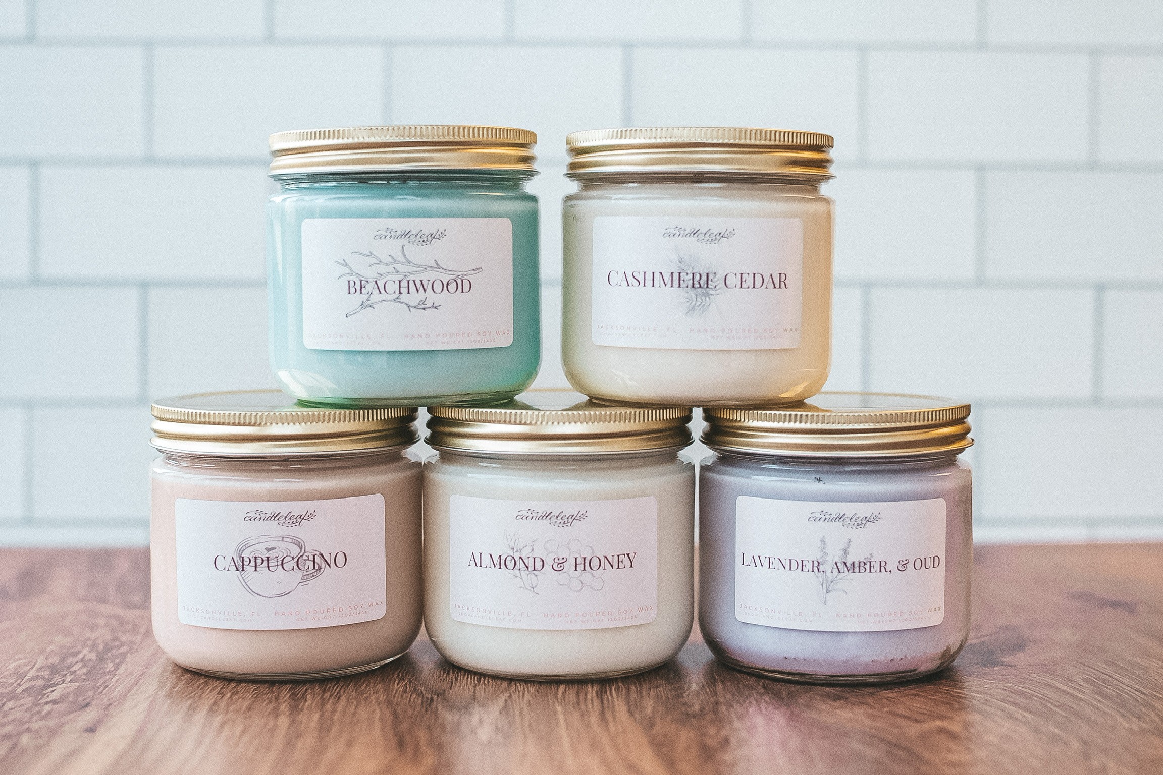 Six candles in glass jars with gold lids arranged on a wooden surface against a white tiled wall.