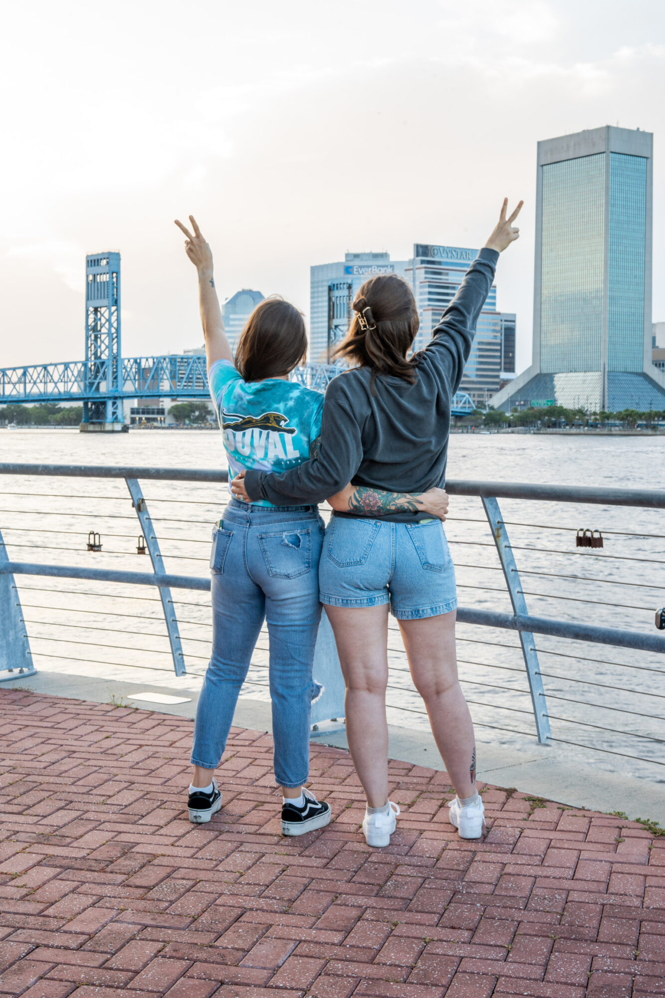 Two women stand by a river, facing away, with arms raised, city skyline in background, overcast sky.