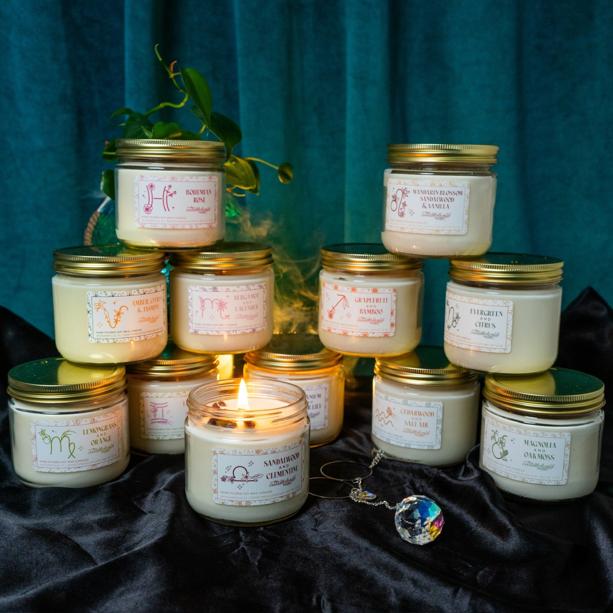 Multiple candles in glass jars with labels, arranged on black fabric with a teal curtain background, some lit, with a small decorative object.