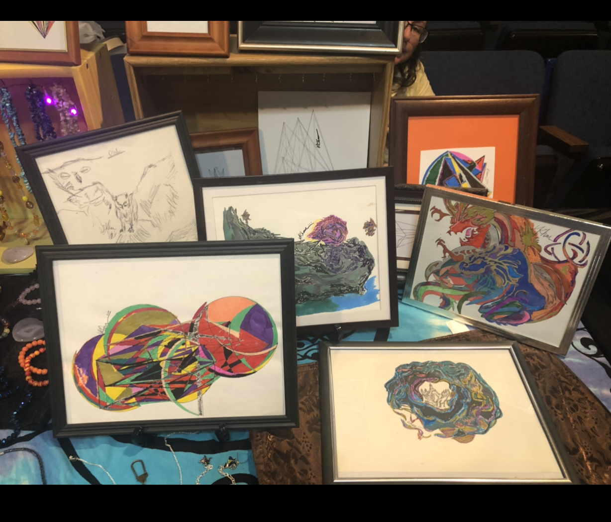 Multiple framed colorful and black-and-white drawings and paintings displayed on a table, with some leaning against a wall.