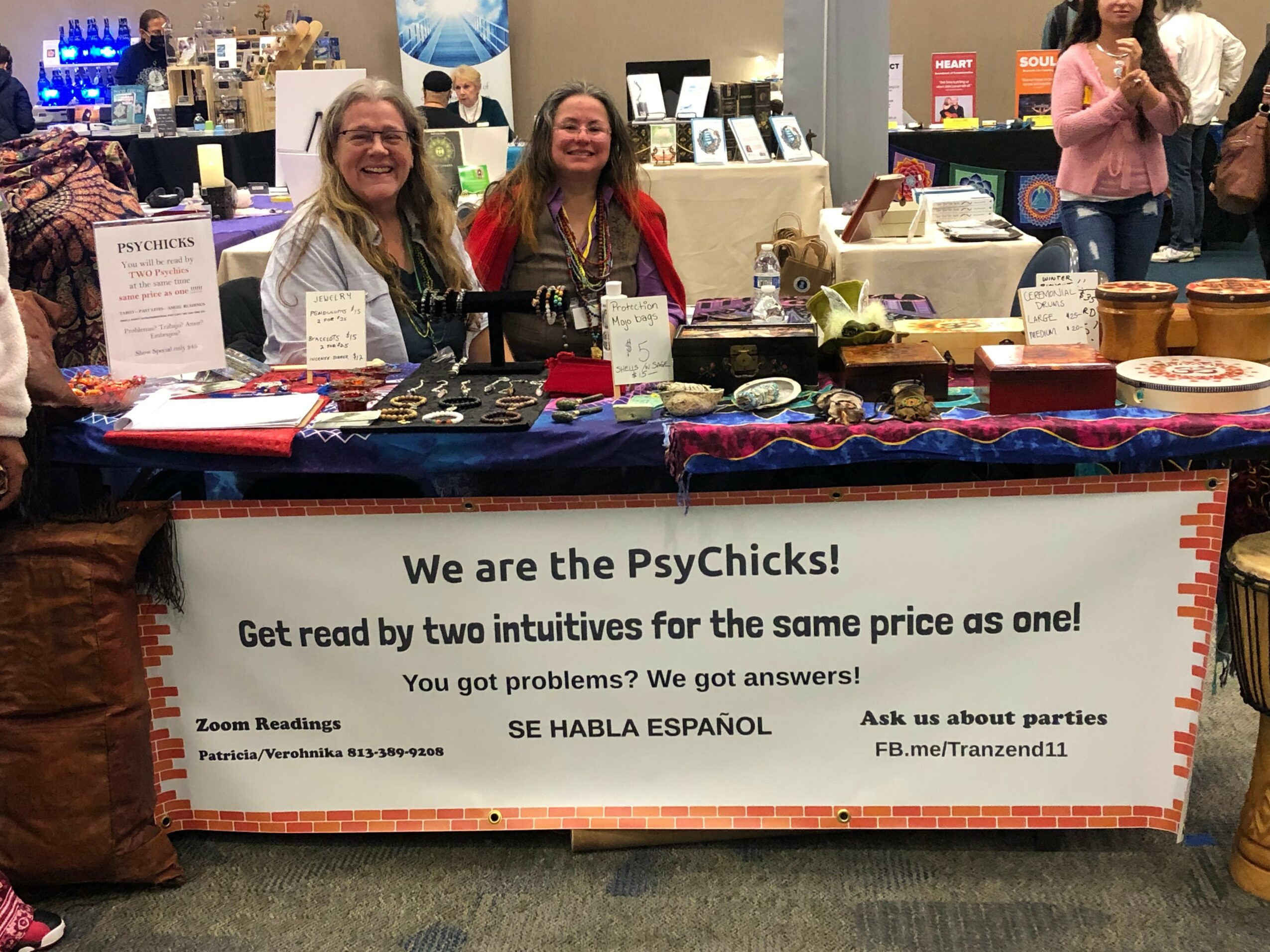 Two women sit behind a table with signs and items at a booth in a busy indoor event space.
