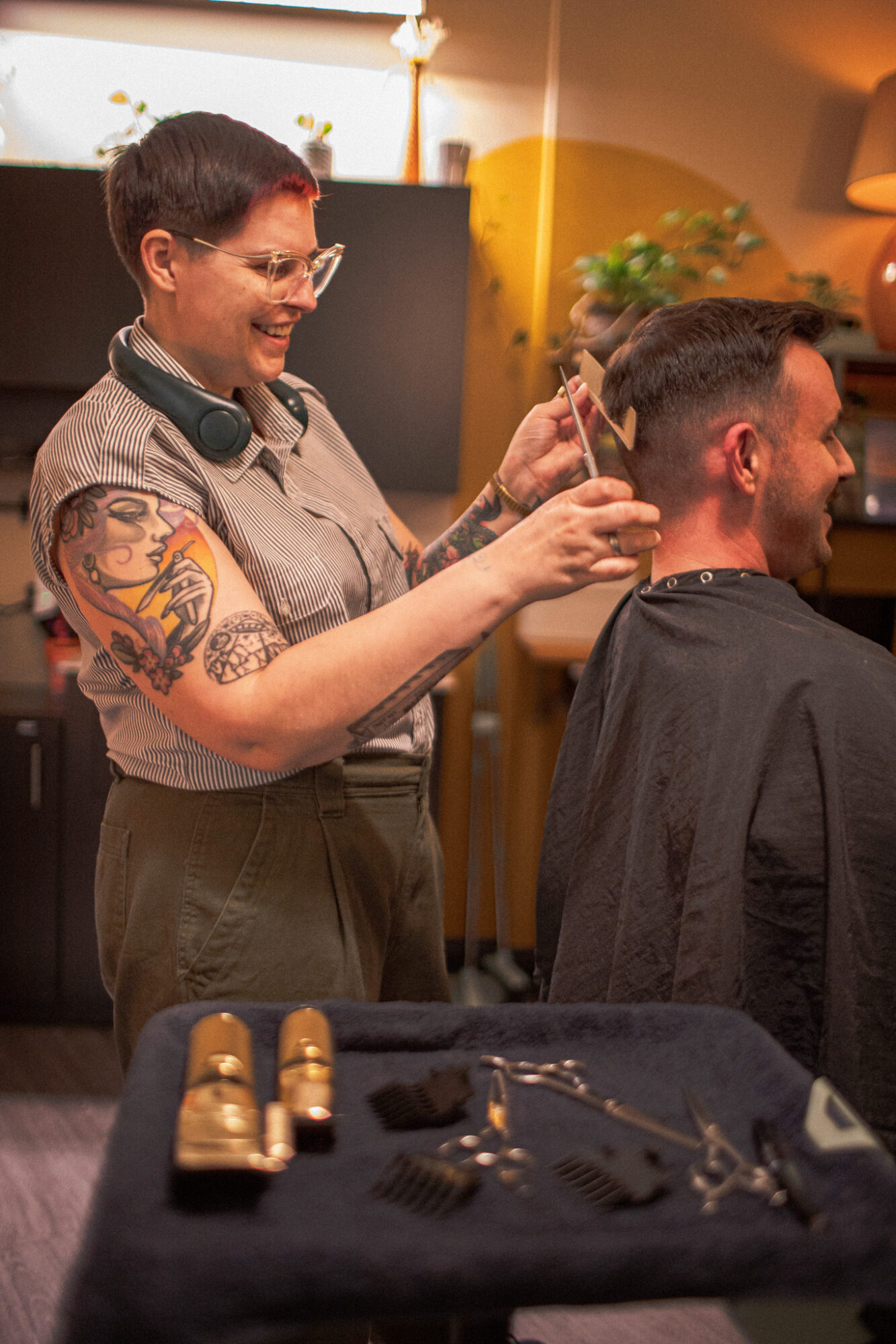 Woman with short hair and glasses cuts a man's hair in a salon, smiling, with a tattoo on her arm, and a black cape on the man.
