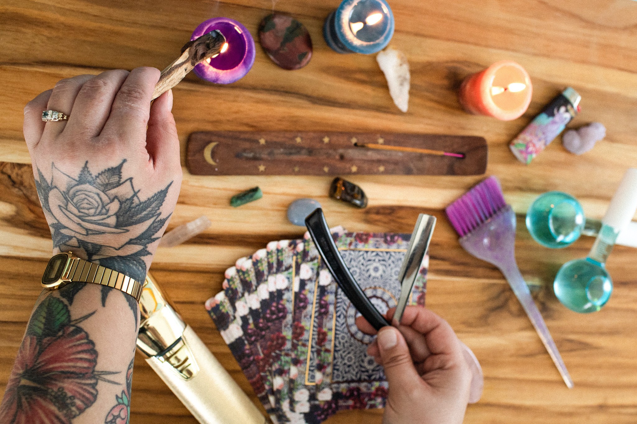 Person's hand with tattoo and ring holding a small purple container, surrounded by tarot cards, candles, and crystals on wooden surface.