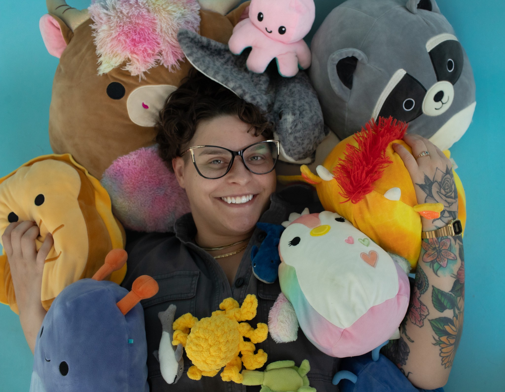 Person with glasses smiling surrounded by colorful plush toys, including animals and fantasy characters, against a blue background.