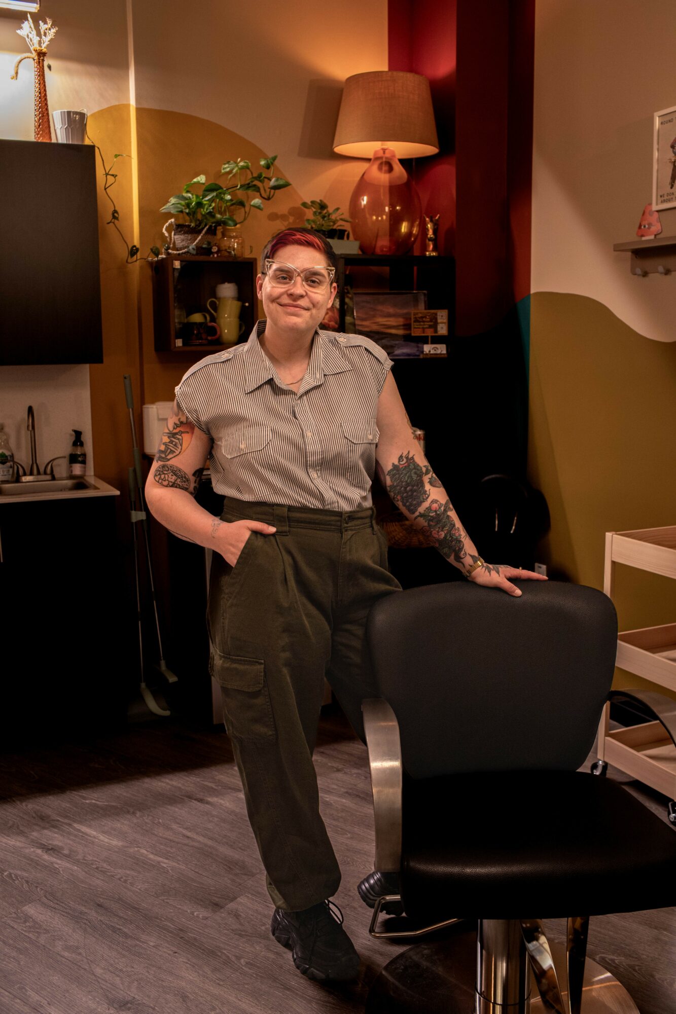 Woman with tattoos, glasses, and short hair standing in a room with warm lighting, a lamp, and plants.