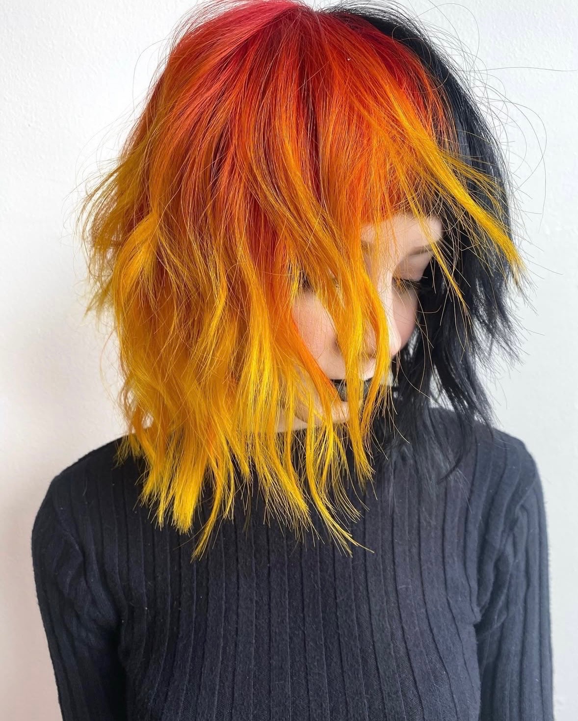 Person with vibrant, multi-colored hair in red, orange, yellow, and black, wearing a dark sweater, facing downward.