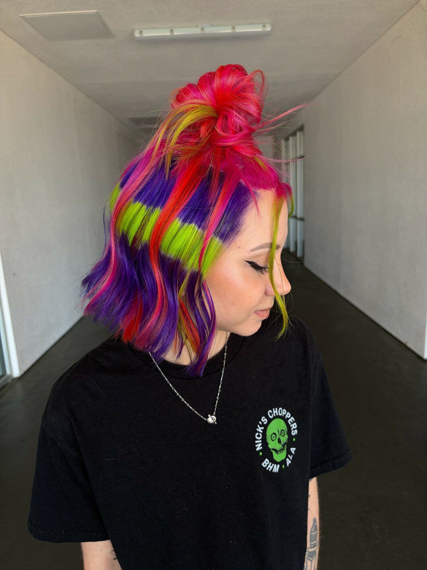 Side profile of a woman with colorful, wavy hair in pink, yellow, green, purple, and blue, wearing a black shirt with a green logo.