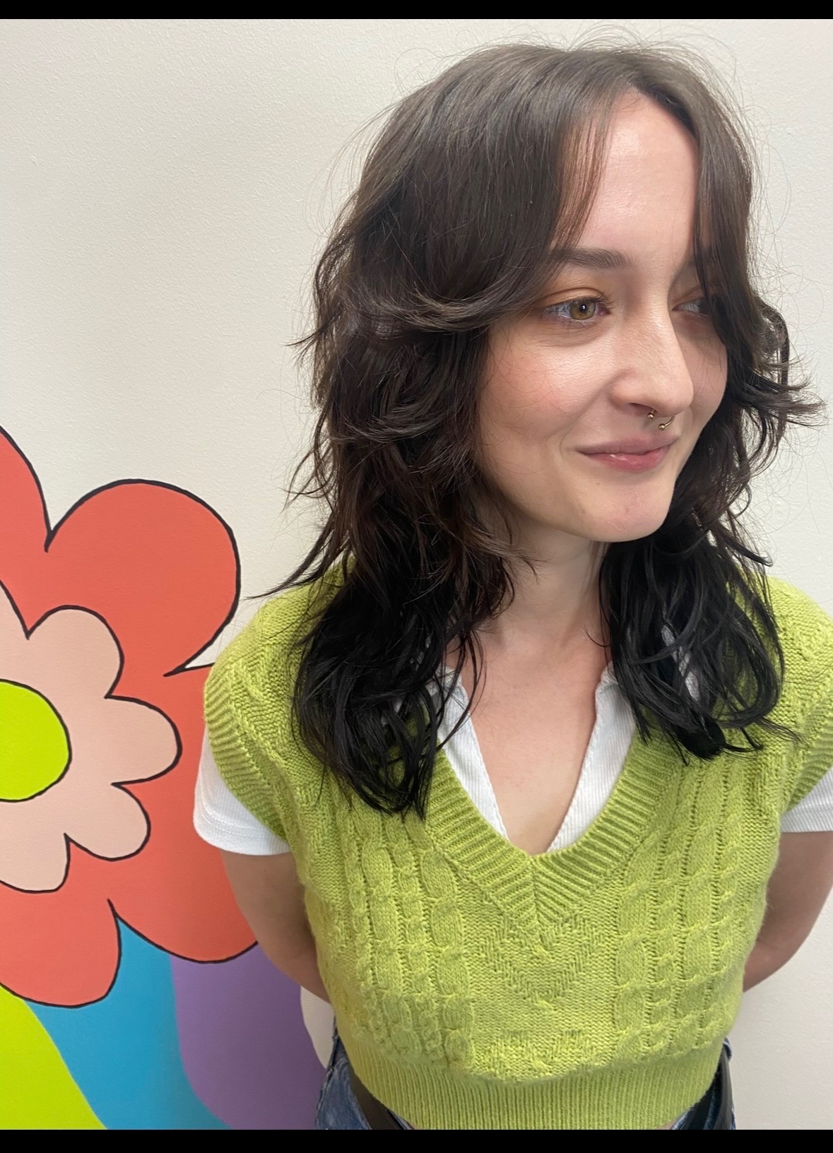 Young woman with wavy dark hair wearing a lime green sweater and white shirt, smiling, standing near a colorful flower illustration.