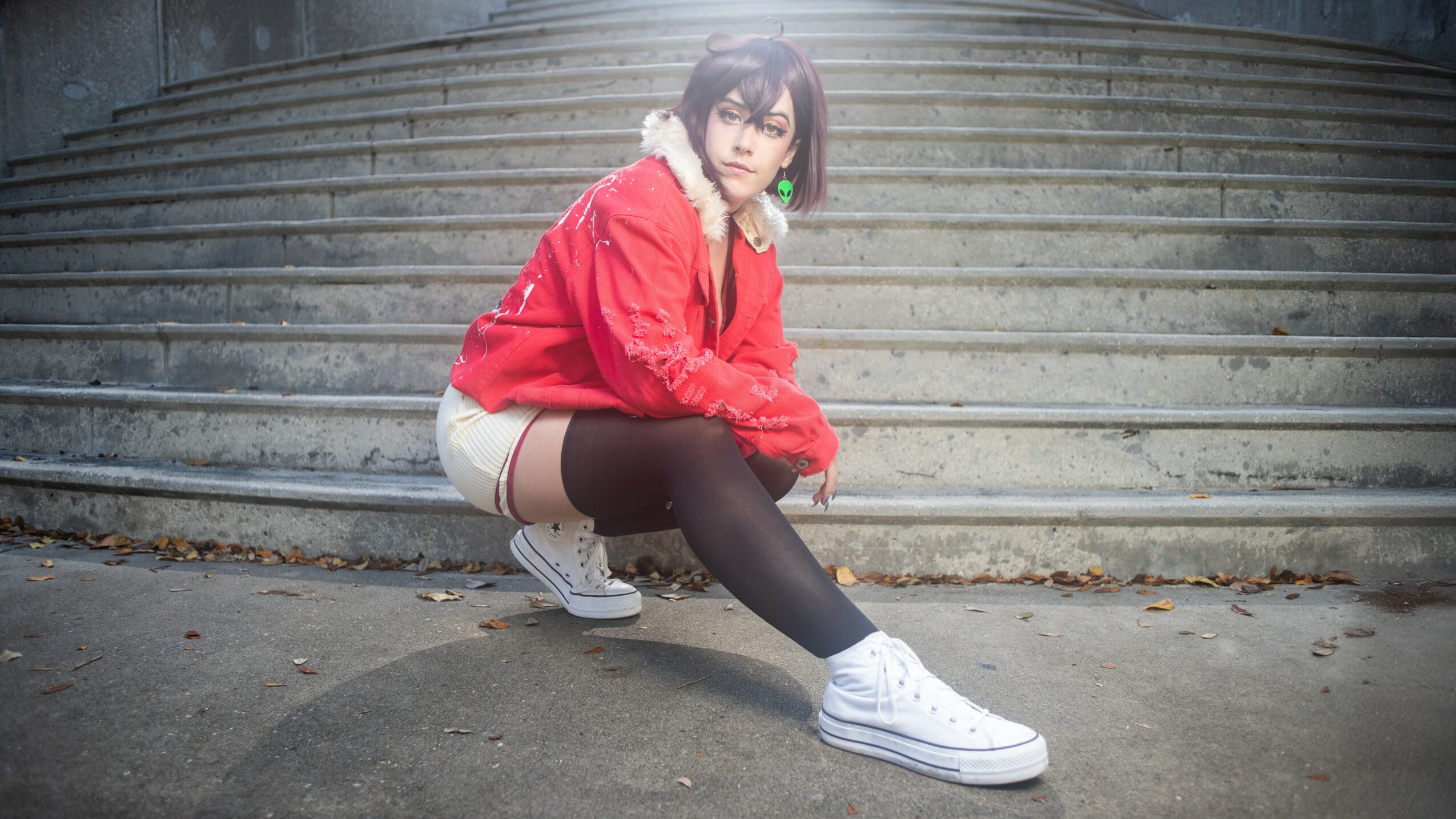 Person crouching on ground in front of concrete stairs, wearing red jacket, shorts, black tights, and white sneakers.