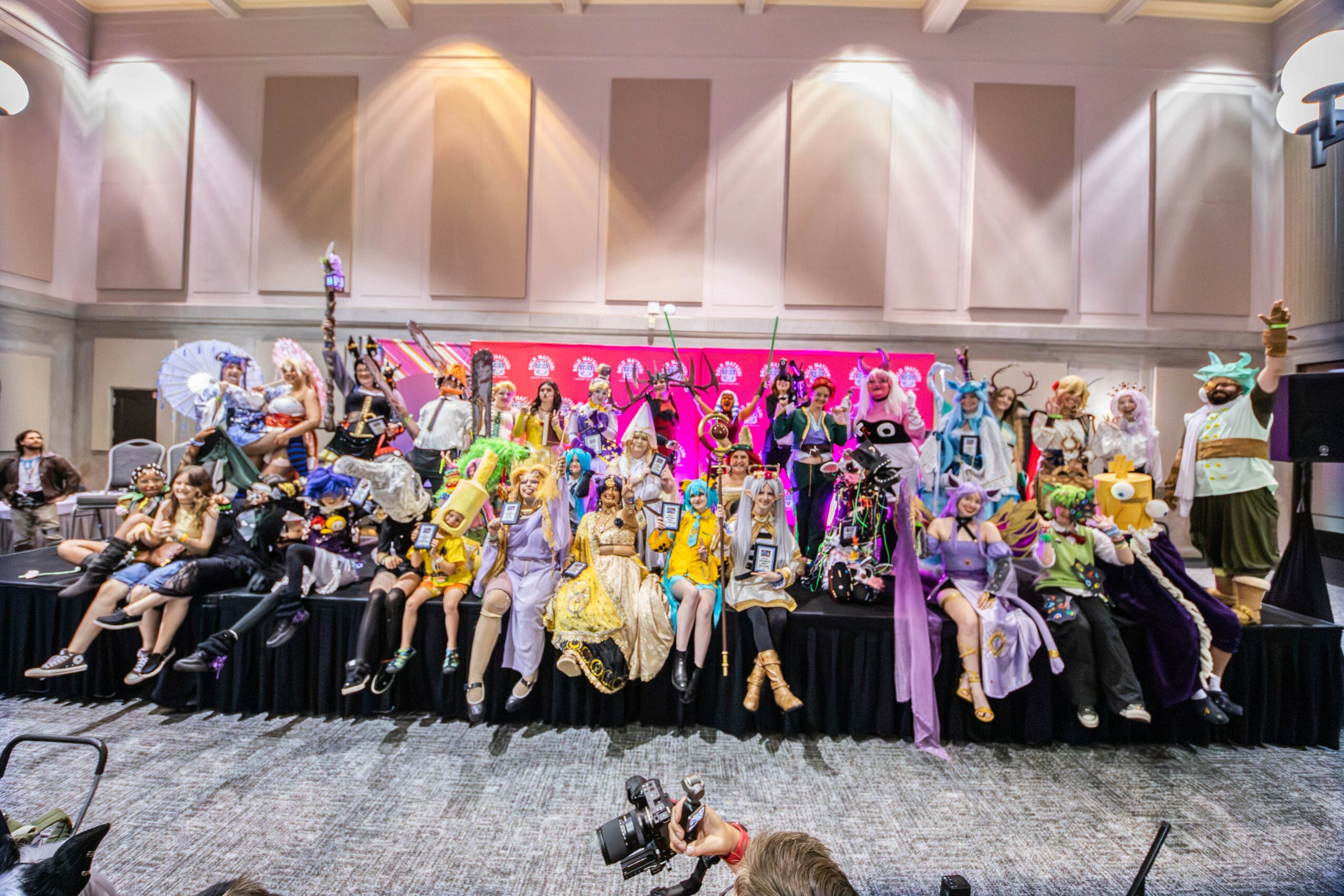 Group of people in costumes and masks on stage with pink backdrop, in a large indoor hall, some seated and some standing.