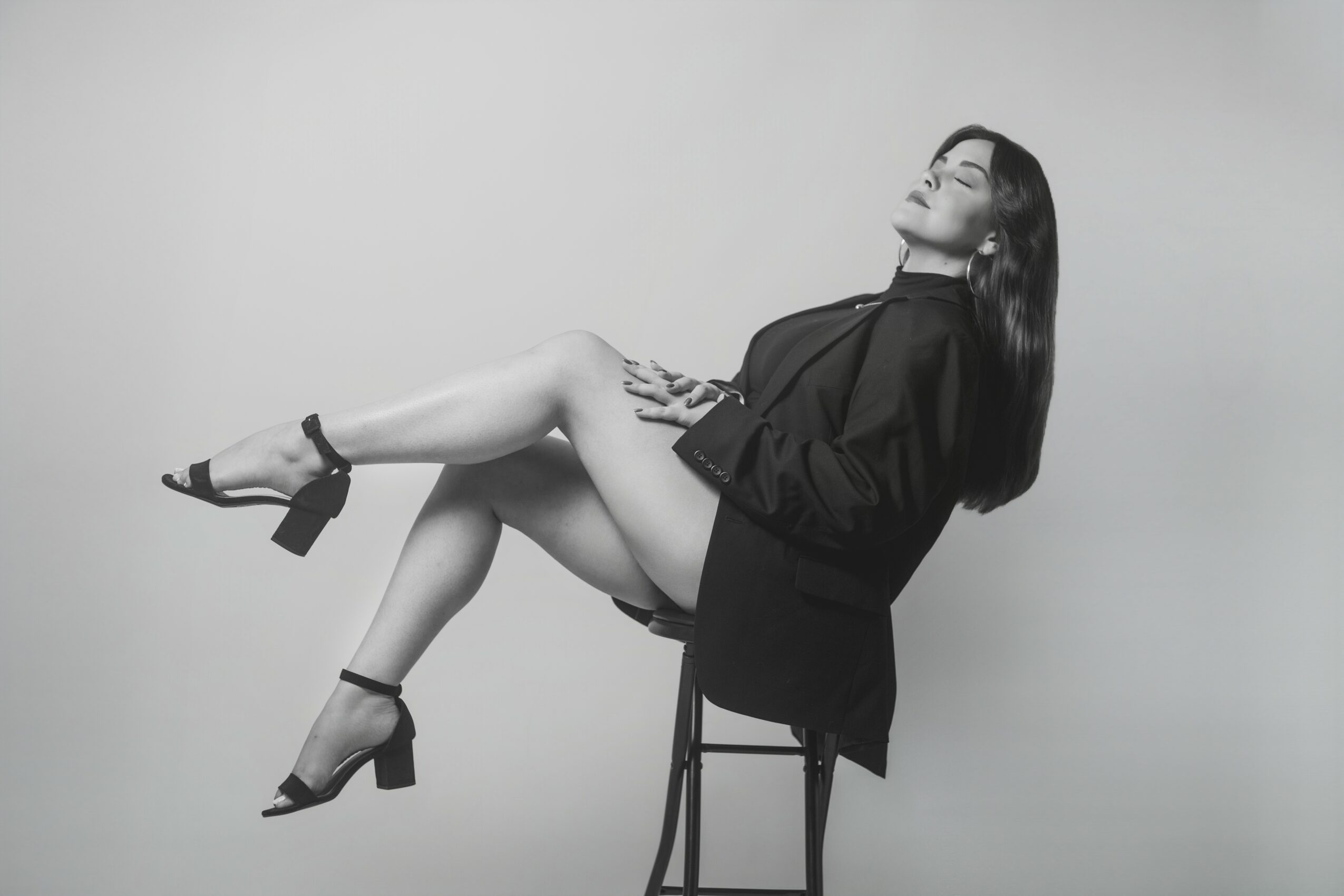 Woman sitting sideways on a chair with legs extended, wearing a blazer and high heels, looking upward.