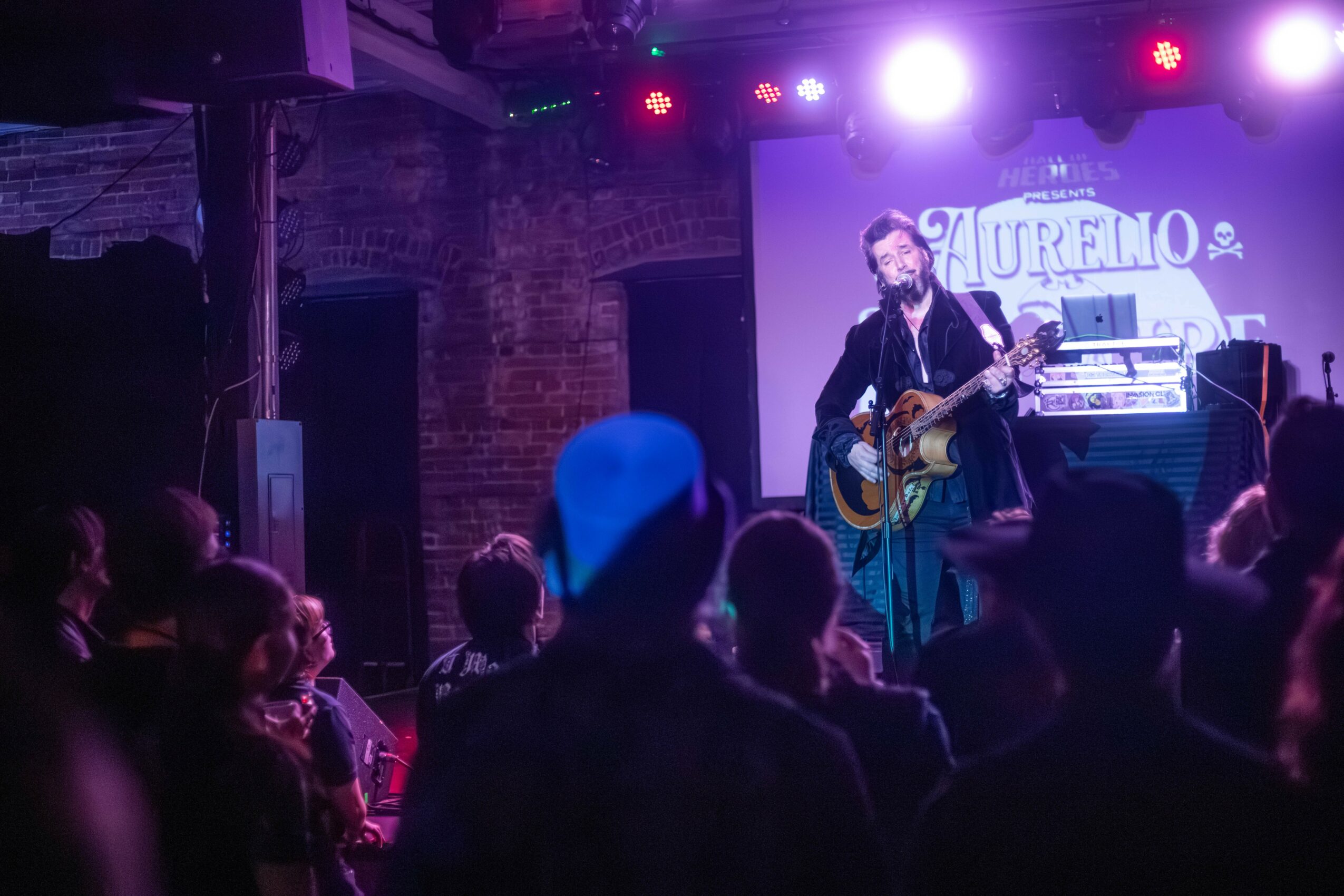 Musician performing on stage with guitar, audience watching, stage lights, and a screen in the background.