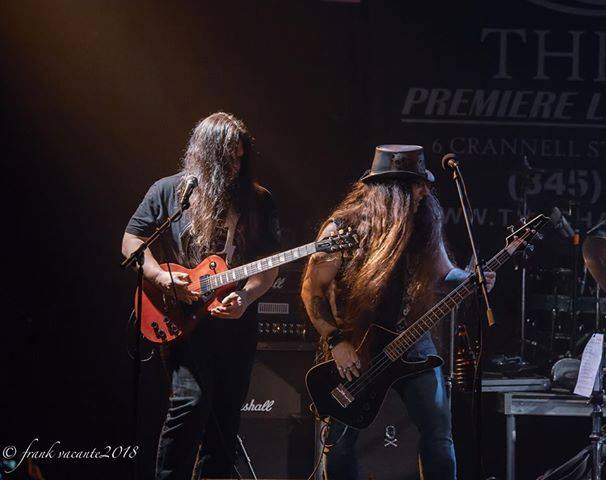 Two musicians playing guitars on stage with dark background and stage lights, one with long hair and a hat, the other with long hair.