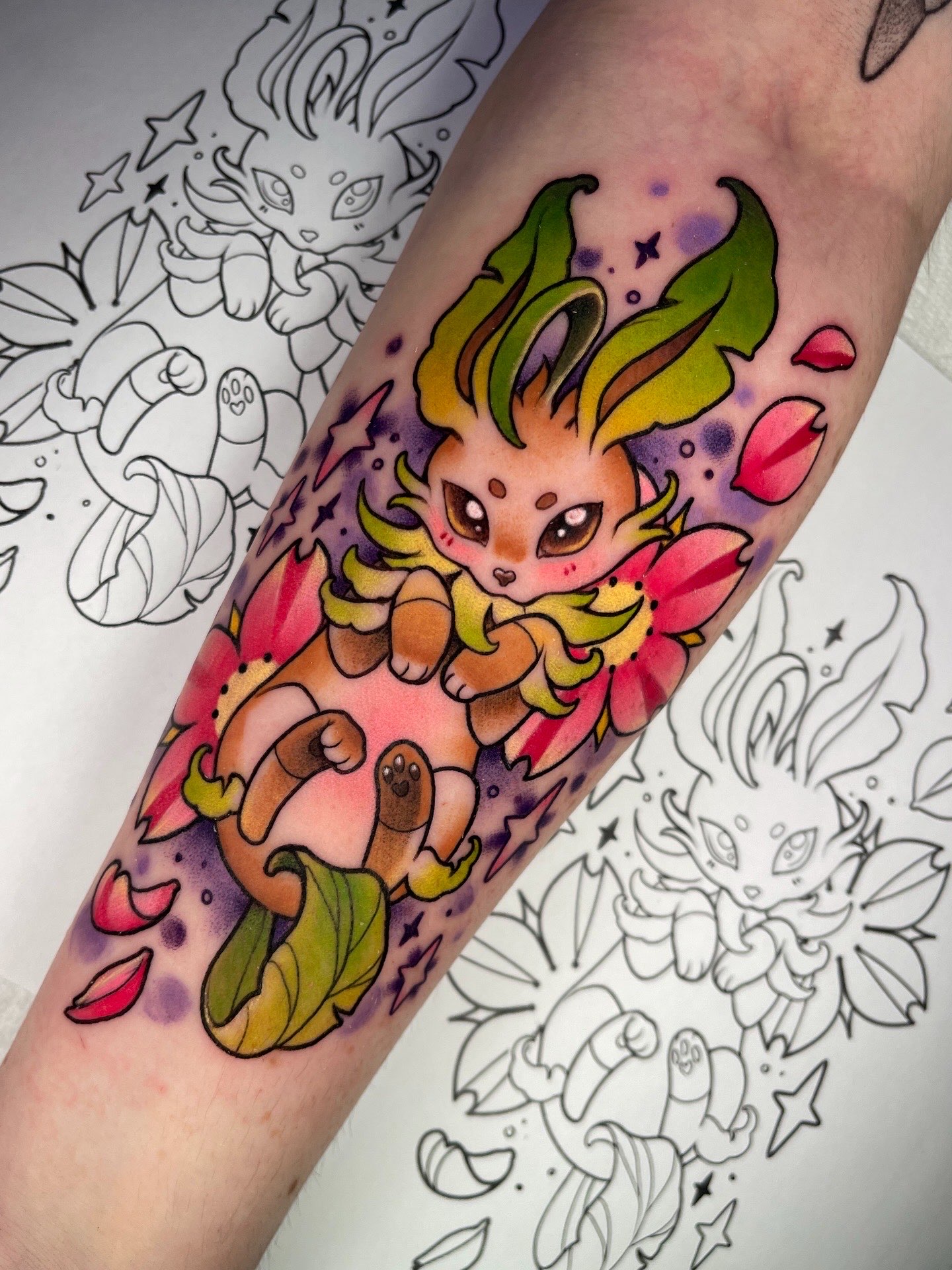 Colorful tattoo of a cute creature with large green ears, surrounded by pink petals and stars, on a person's arm.