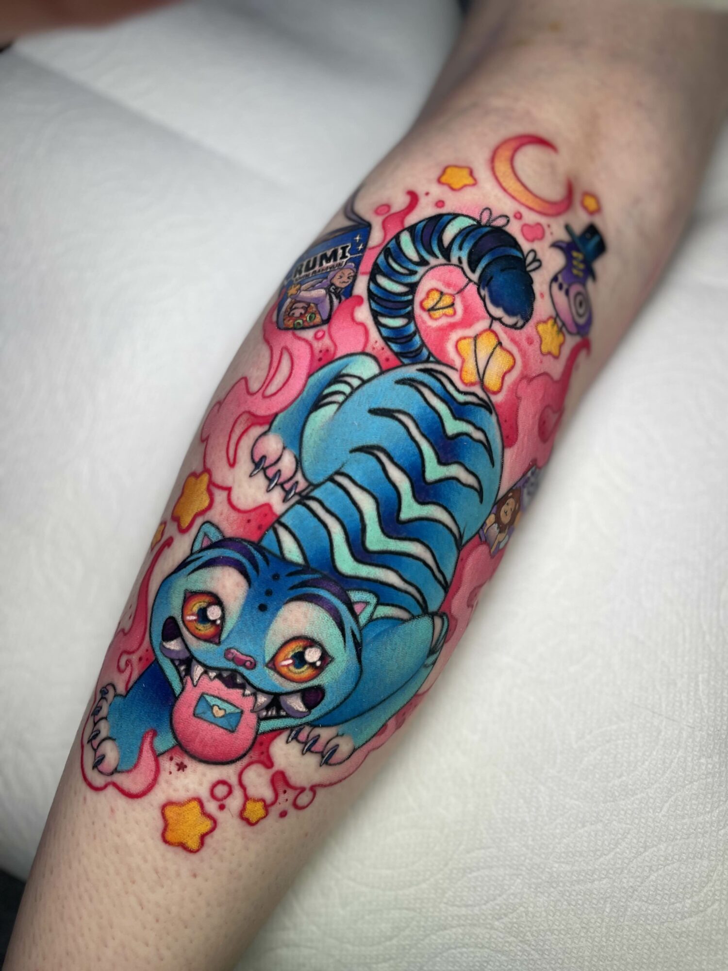 Colorful tattoo of a blue tiger with pink accents, surrounded by stars, moons, and clouds on a person's arm.