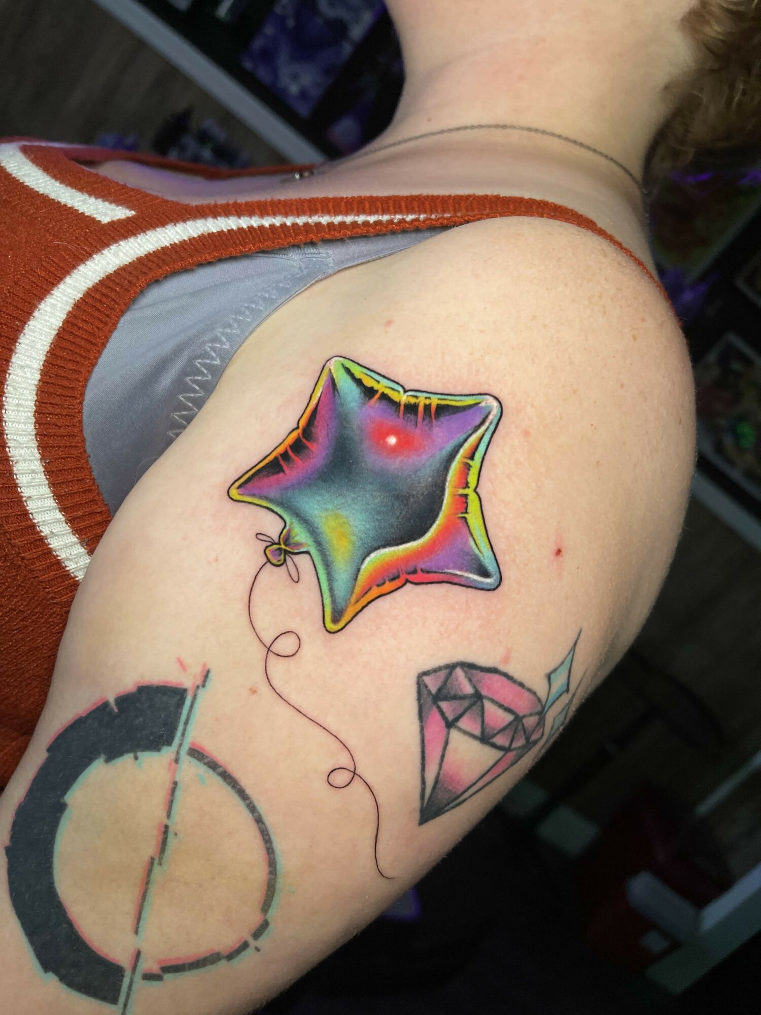 Colorful star-shaped tattoo with a rainbow gradient on a person's shoulder, along with a pink diamond and a partial circle tattoo.