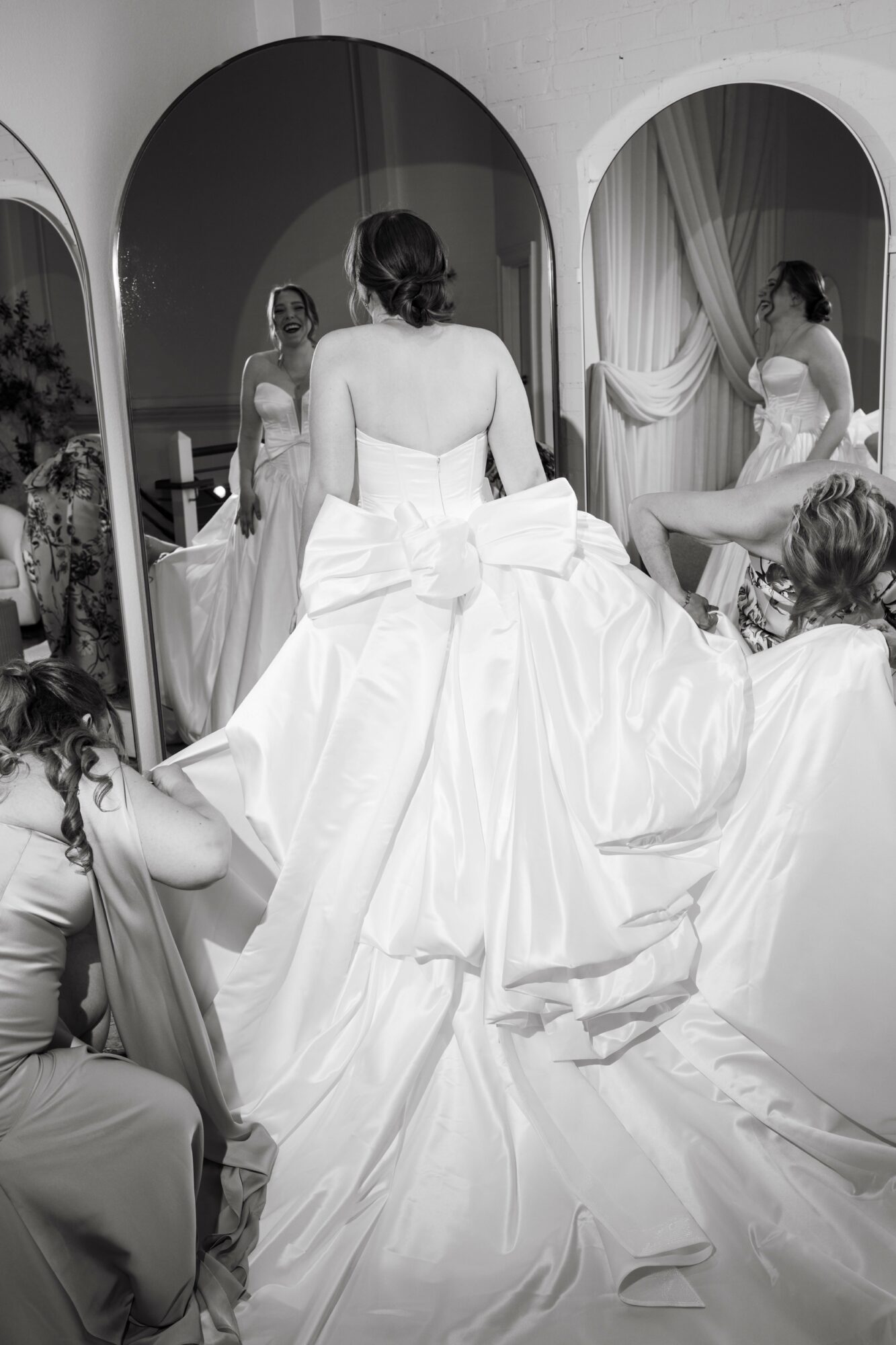 Group of women in wedding dresses preparing in front of mirrors, with one woman adjusting her gown, in a room with arched mirrors.