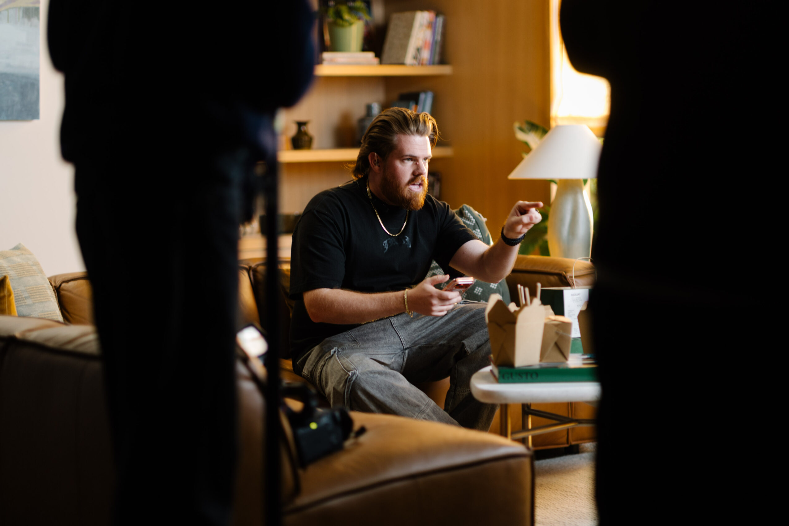 Man with glasses and beard sitting on a couch, holding a card, in a cozy room with books and a lamp